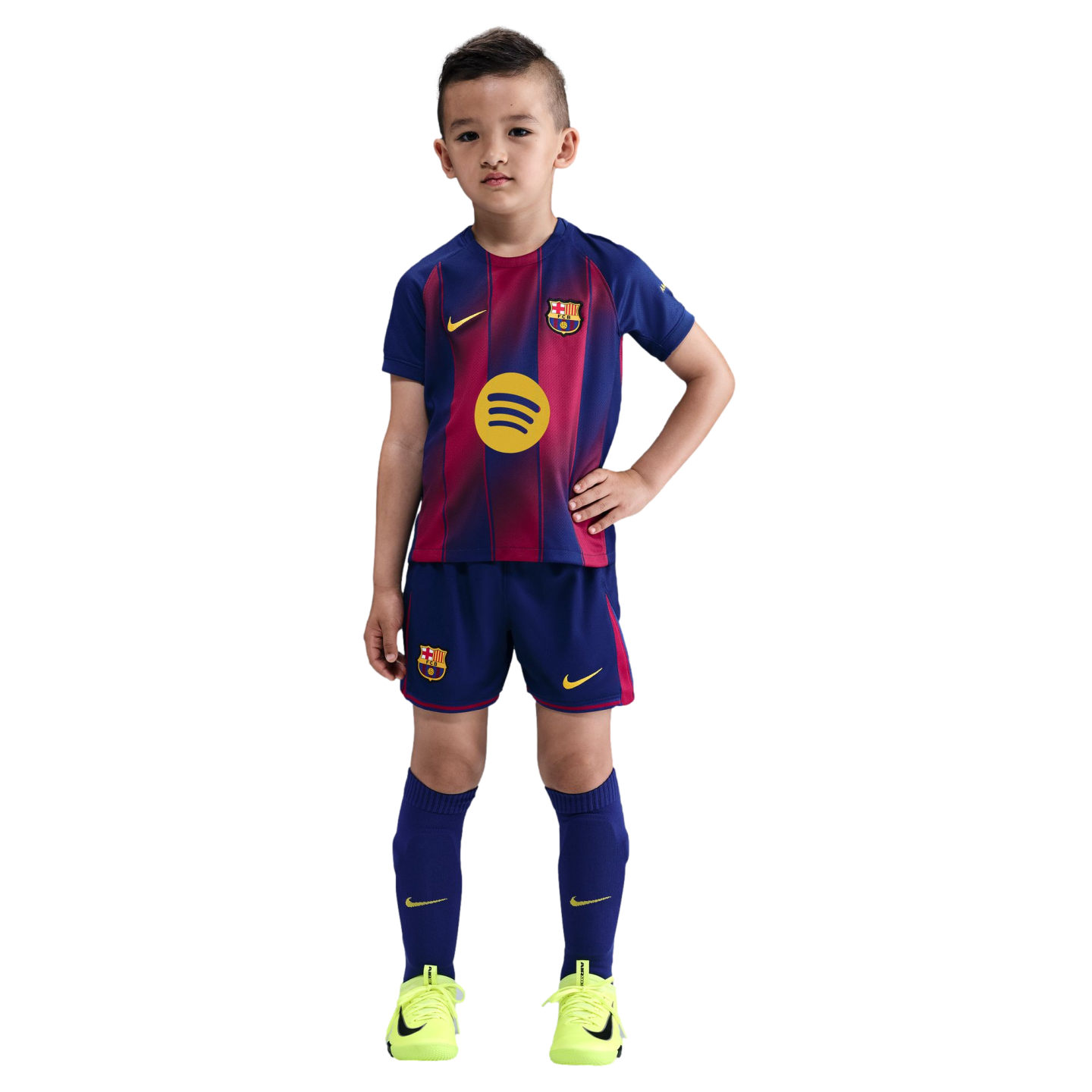 A young boy poses in a full FC Barcelona soccer kit with neon yellow cleats against a black background.