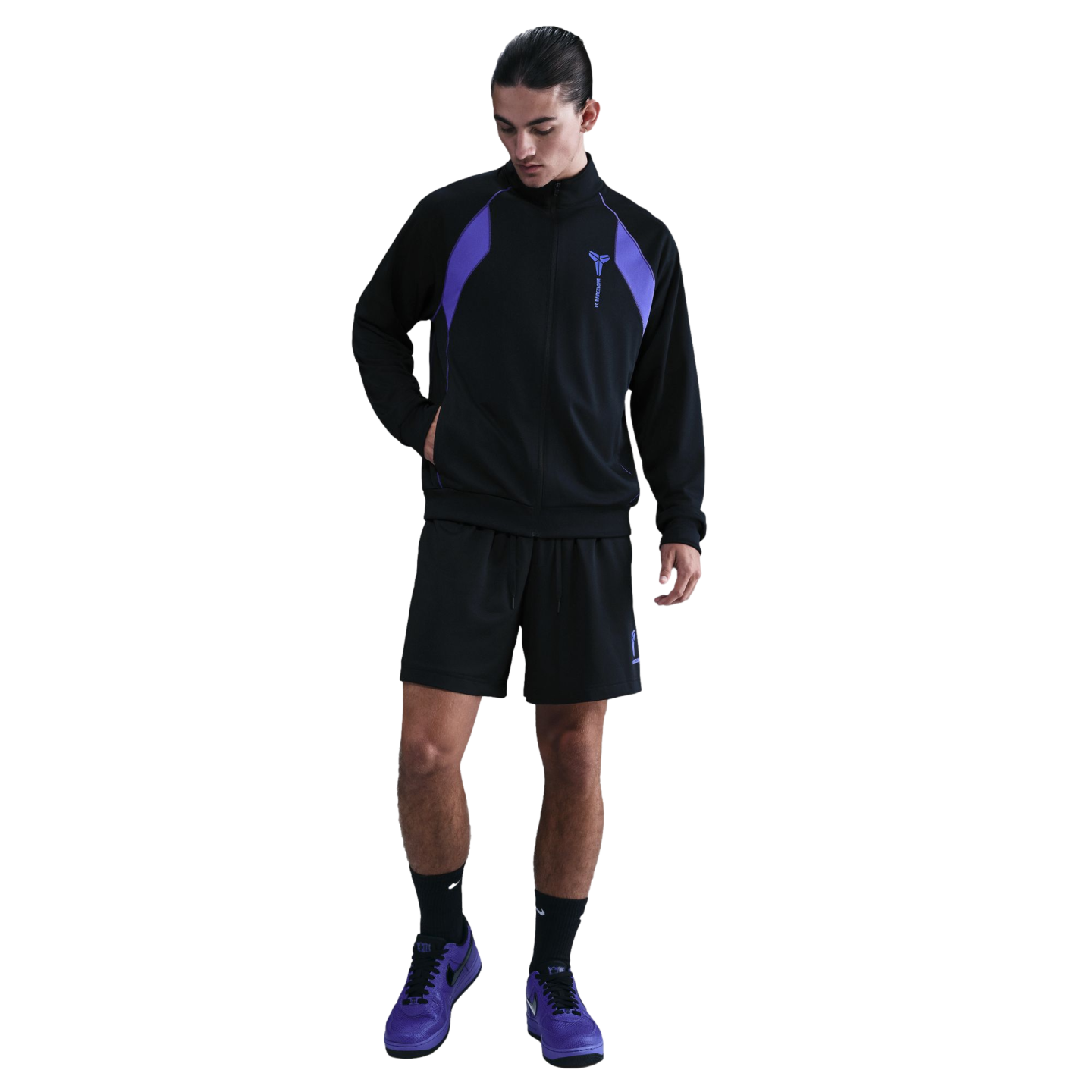 A man wearing a black tracksuit jacket with purple shoulder panels, black shorts, and purple sneakers against a solid black background.