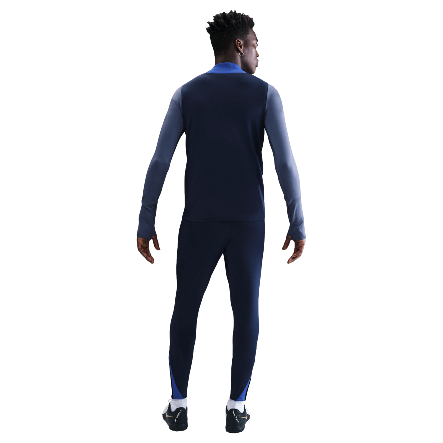 Rear view of a man wearing a navy blue and light blue long-sleeved athletic top and matching pants with black soccer cleats against a white background.