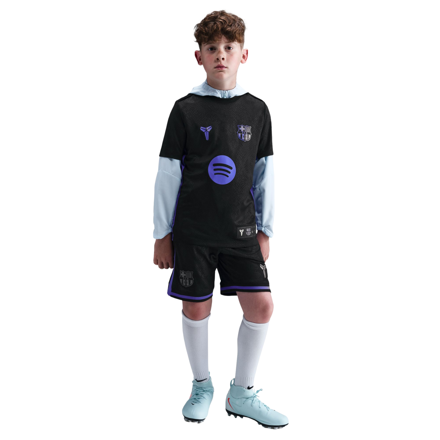 A young boy models a black FC Barcelona soccer kit with purple accents and logos, worn over a light blue long-sleeved shirt with matching light blue cleats.
