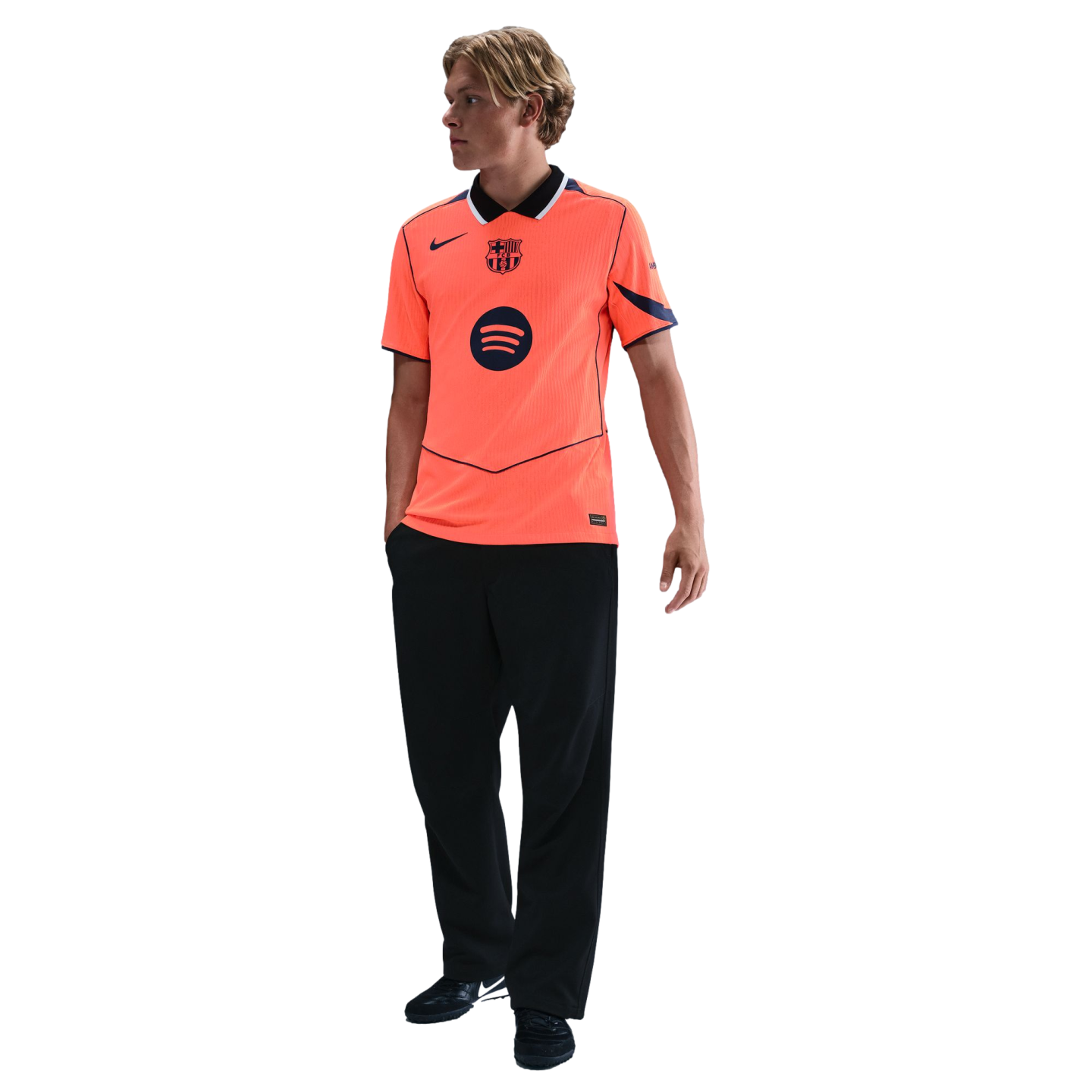 A young blonde man wearing a bright orange FC Barcelona Nike soccer jersey and black trousers, standing against a solid black background.
