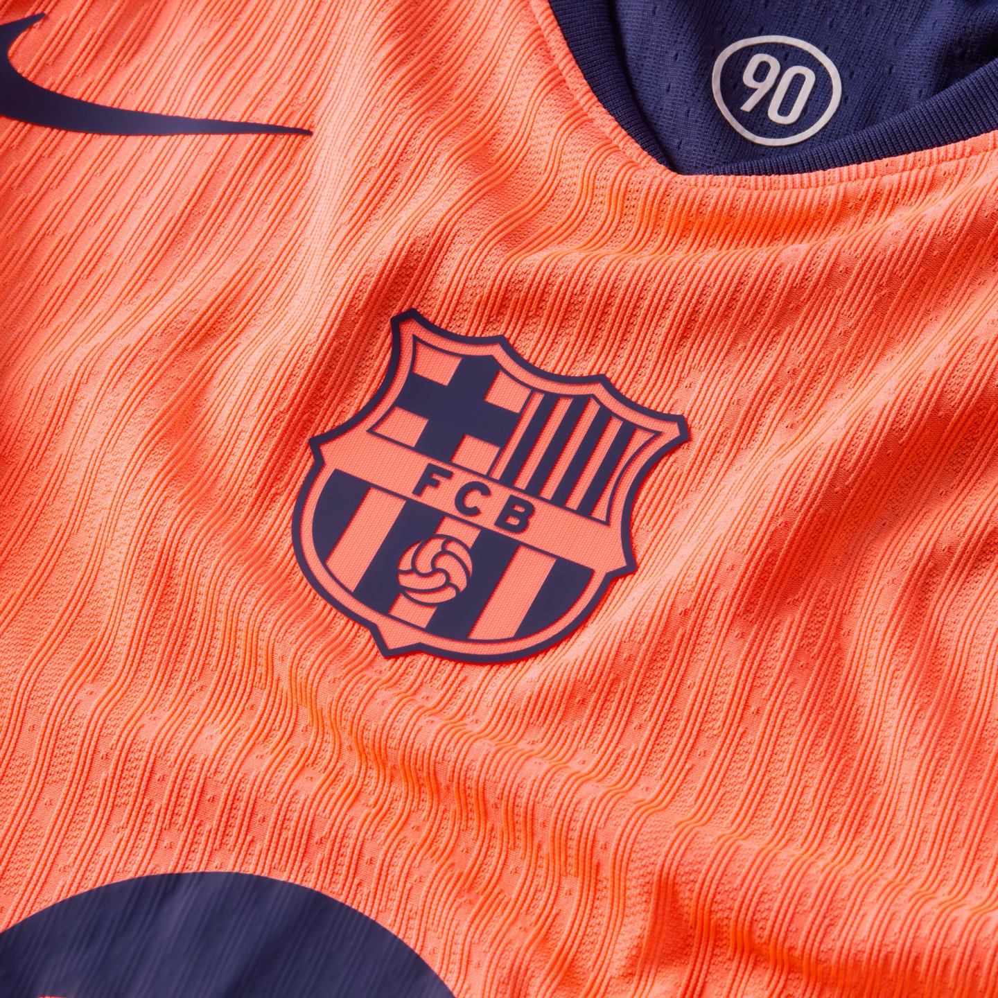 Close-up of an orange FC Barcelona soccer jersey with ribbed texture, featuring the club crest and a "90" logo on the inner collar.