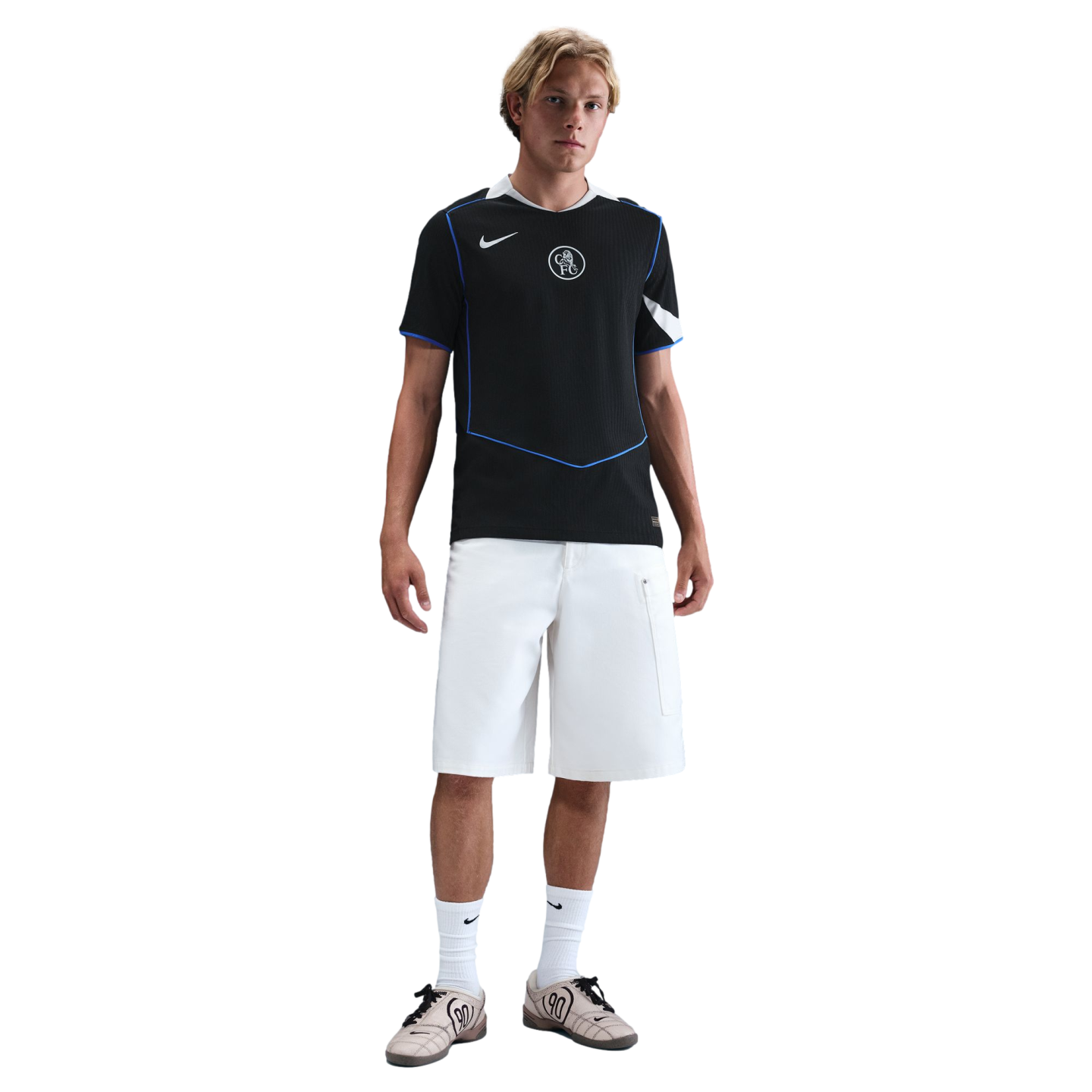 A blonde man stands against a black background wearing a black sports jersey with blue trim, white shorts, white socks, and tan sneakers.