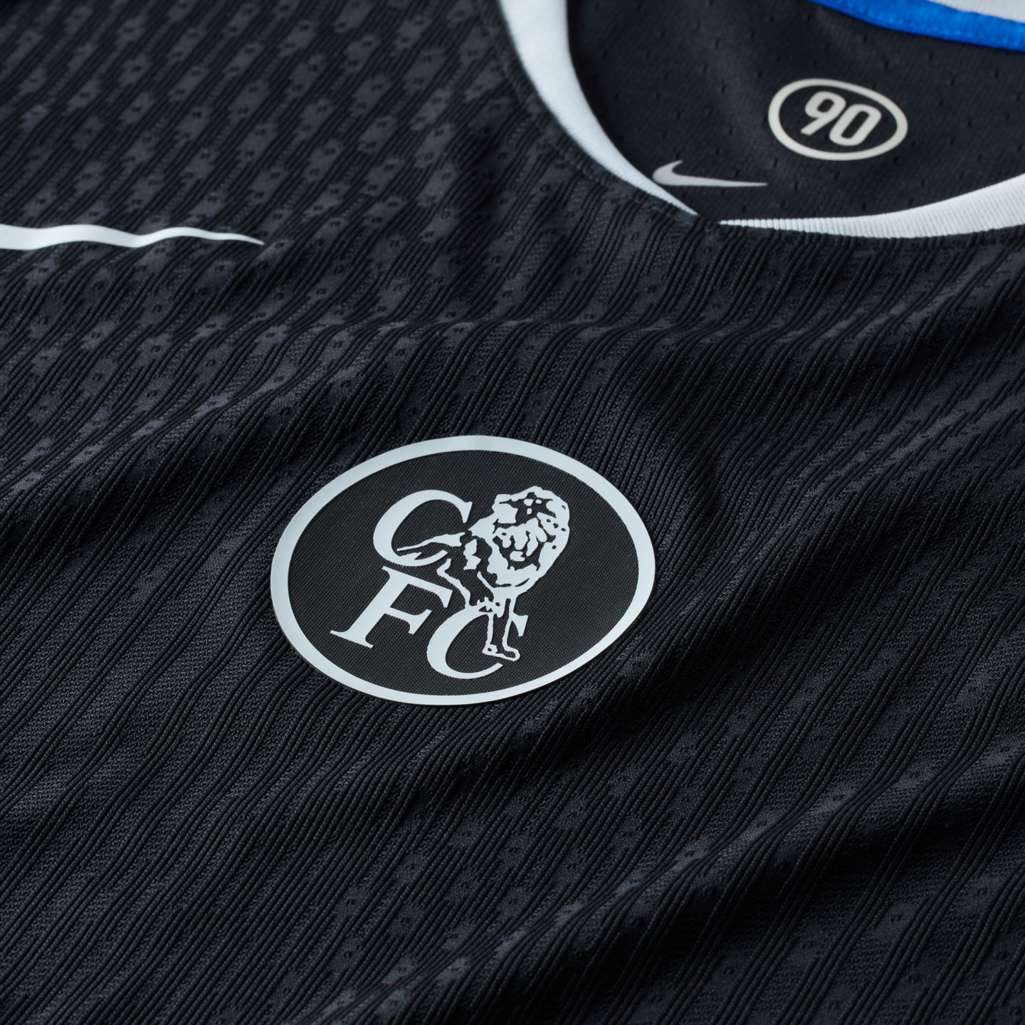 Close-up of a black textured football jersey featuring a white circular Chelsea FC crest, a Nike swoosh, and a "90" logo on the inner collar.