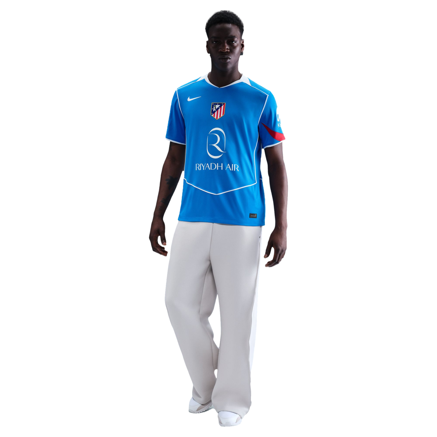 Man in a blue Atletico Madrid soccer jersey and white wide-leg trousers against a black background.