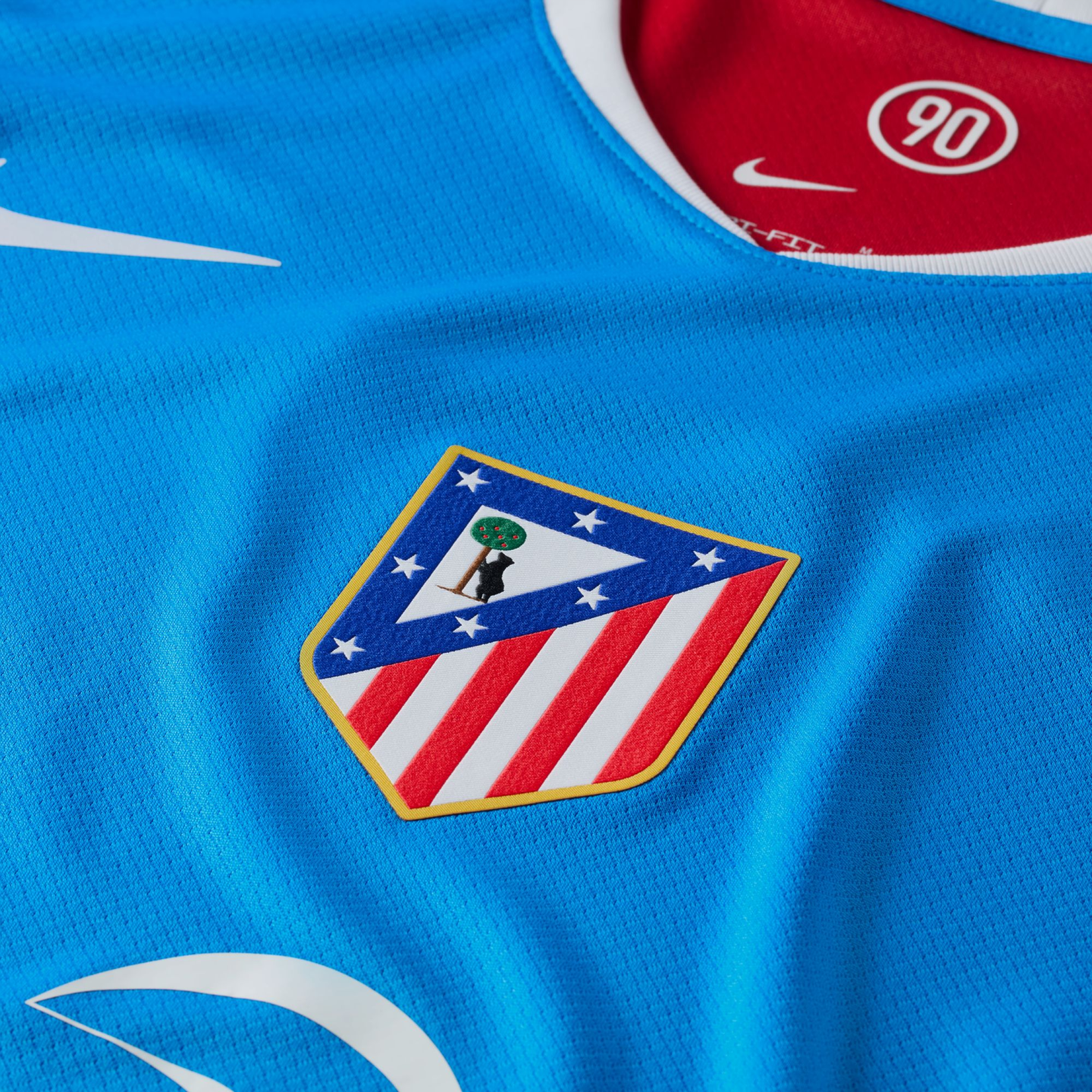 A close-up view of a blue Nike soccer jersey featuring the Atlético Madrid team crest and a red inner collar with a "90" logo.