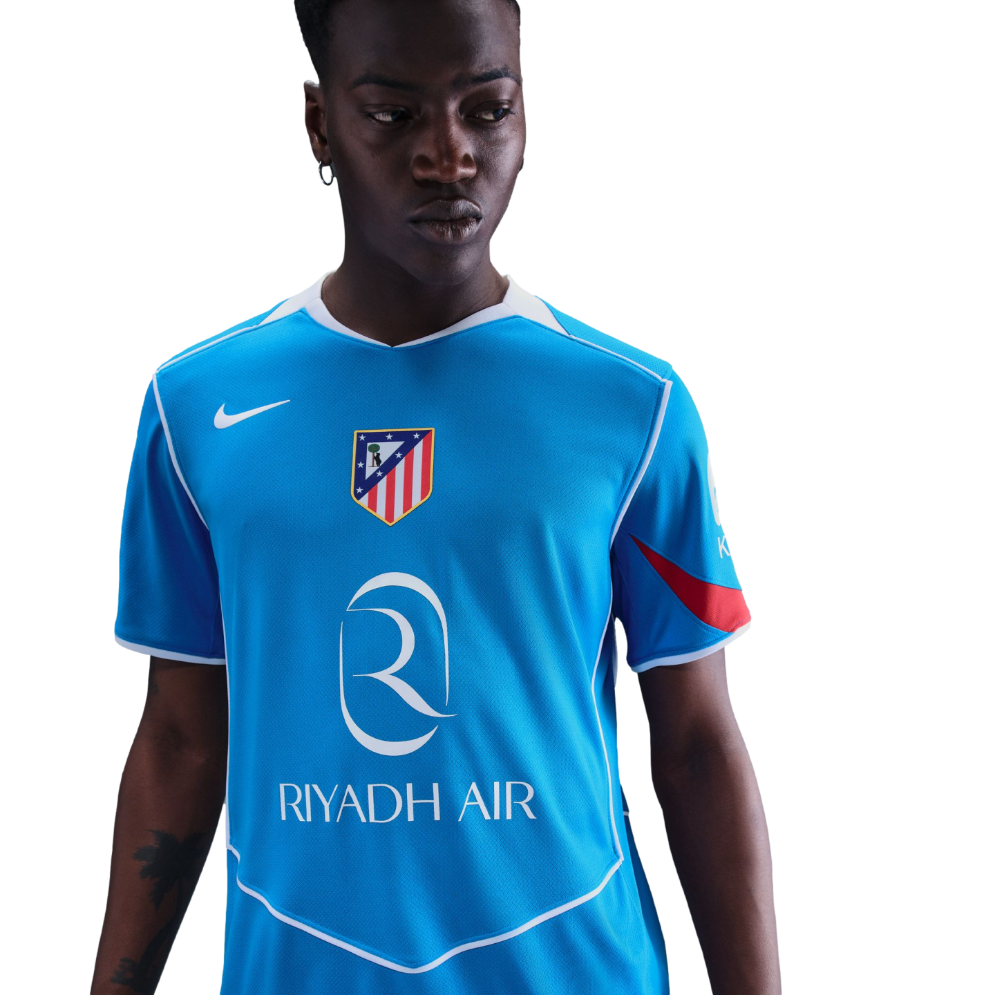 A man with a serious expression wears a light blue Nike soccer jersey with the Atletico Madrid crest and Riyadh Air logo, set against a solid black background.