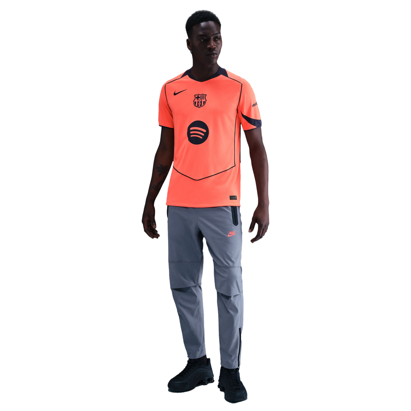 A man modeling a bright orange Nike FC Barcelona jersey, grey athletic pants, and black sneakers against a black background.