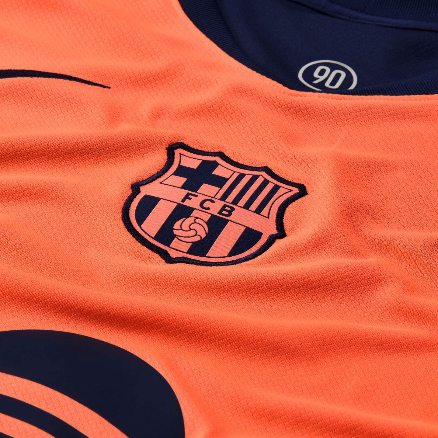 Close-up of a vibrant orange FC Barcelona jersey featuring the club's crest, a navy collar with a "90" logo, and a partial sponsor logo.