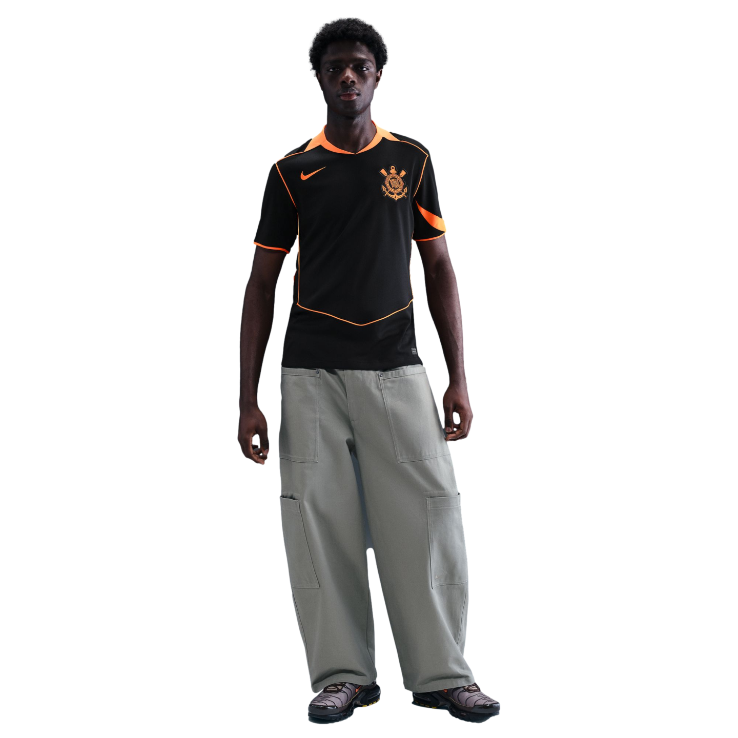 Man wearing a black Nike soccer jersey with orange trim, baggy grey cargo pants, and sneakers, standing against a solid black background.