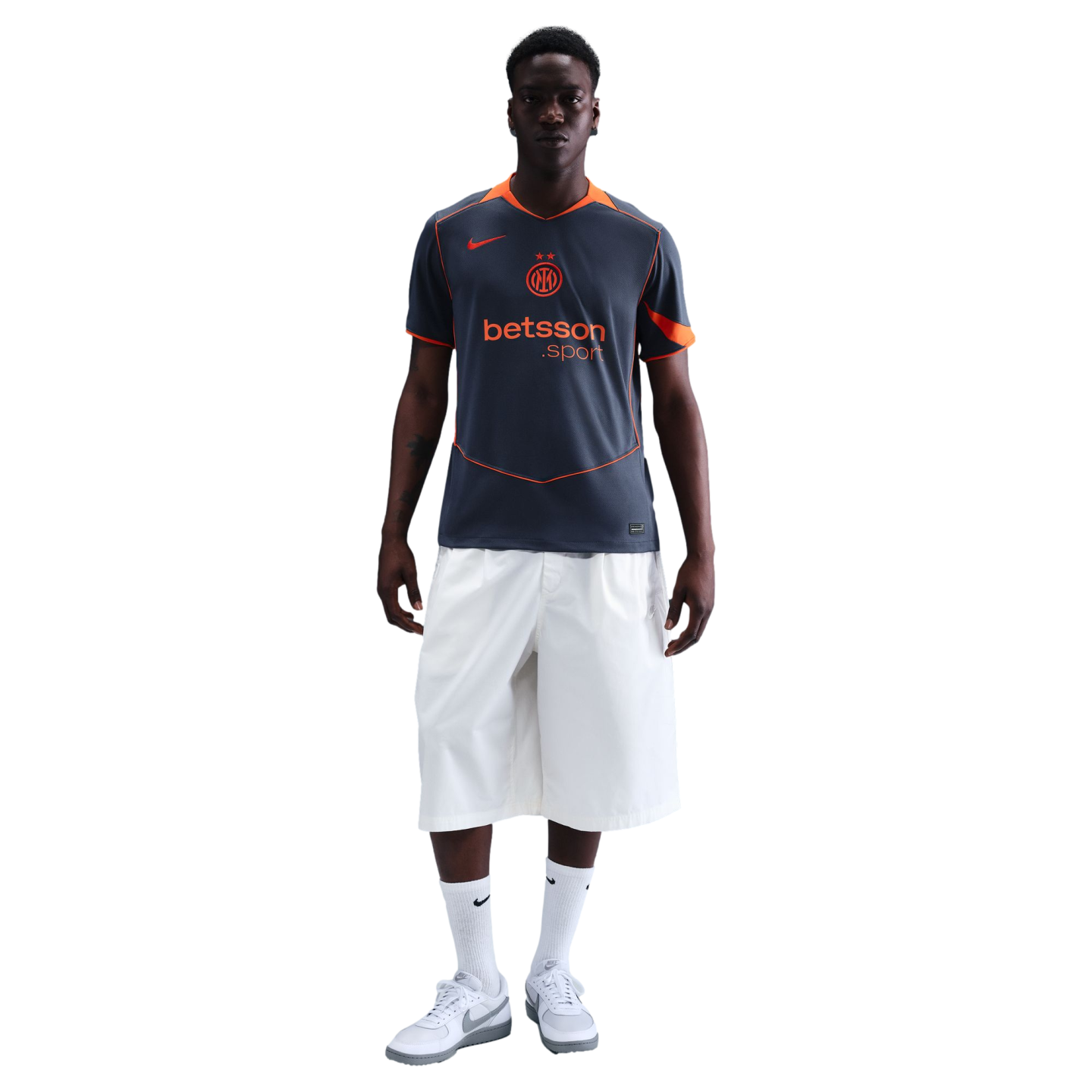 A man wearing a grey and orange Inter Milan training jersey, white knee-length shorts, and white sneakers, standing against a solid black background.