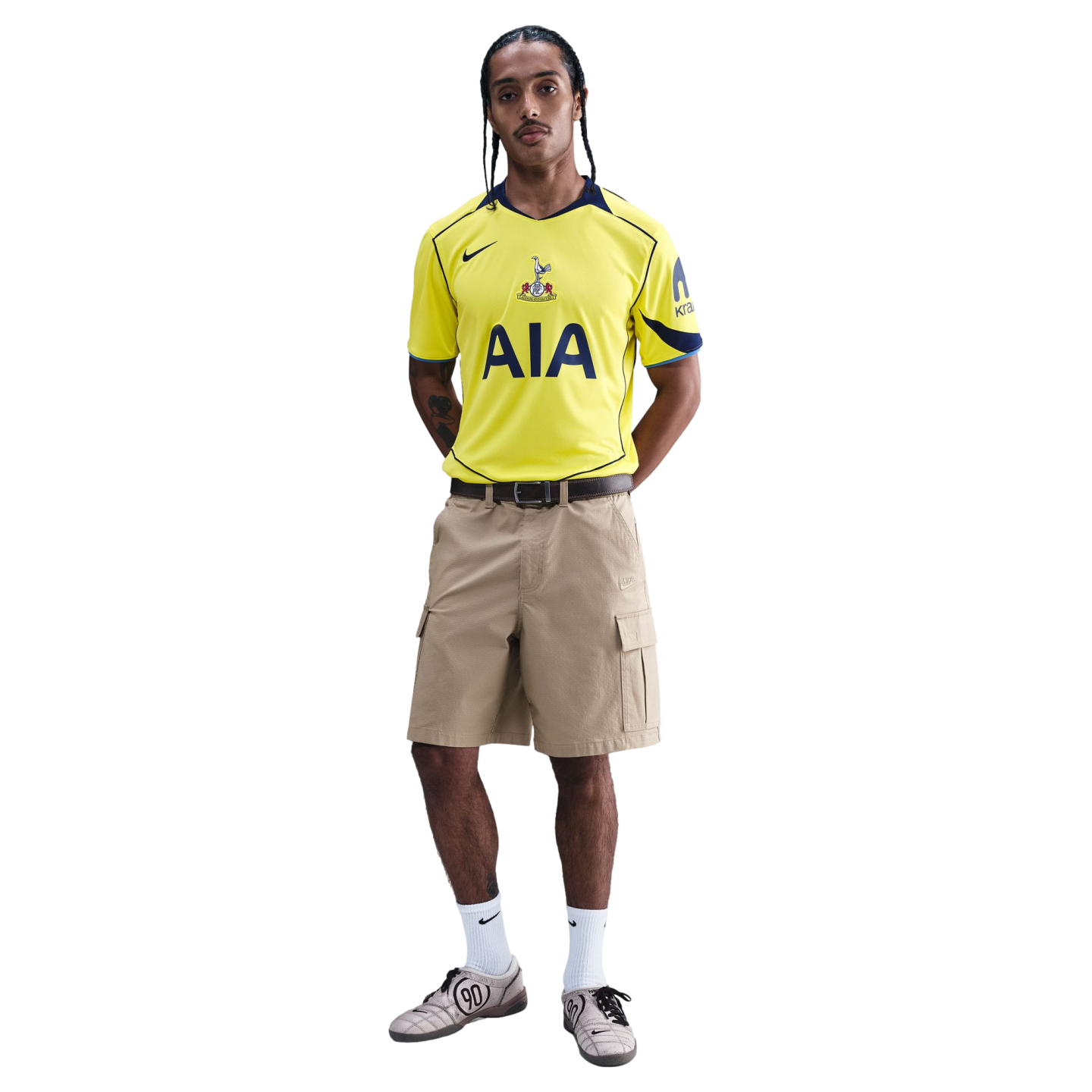 A man models a yellow Tottenham Hotspur football jersey, tan cargo shorts, and grey sneakers against a black background.