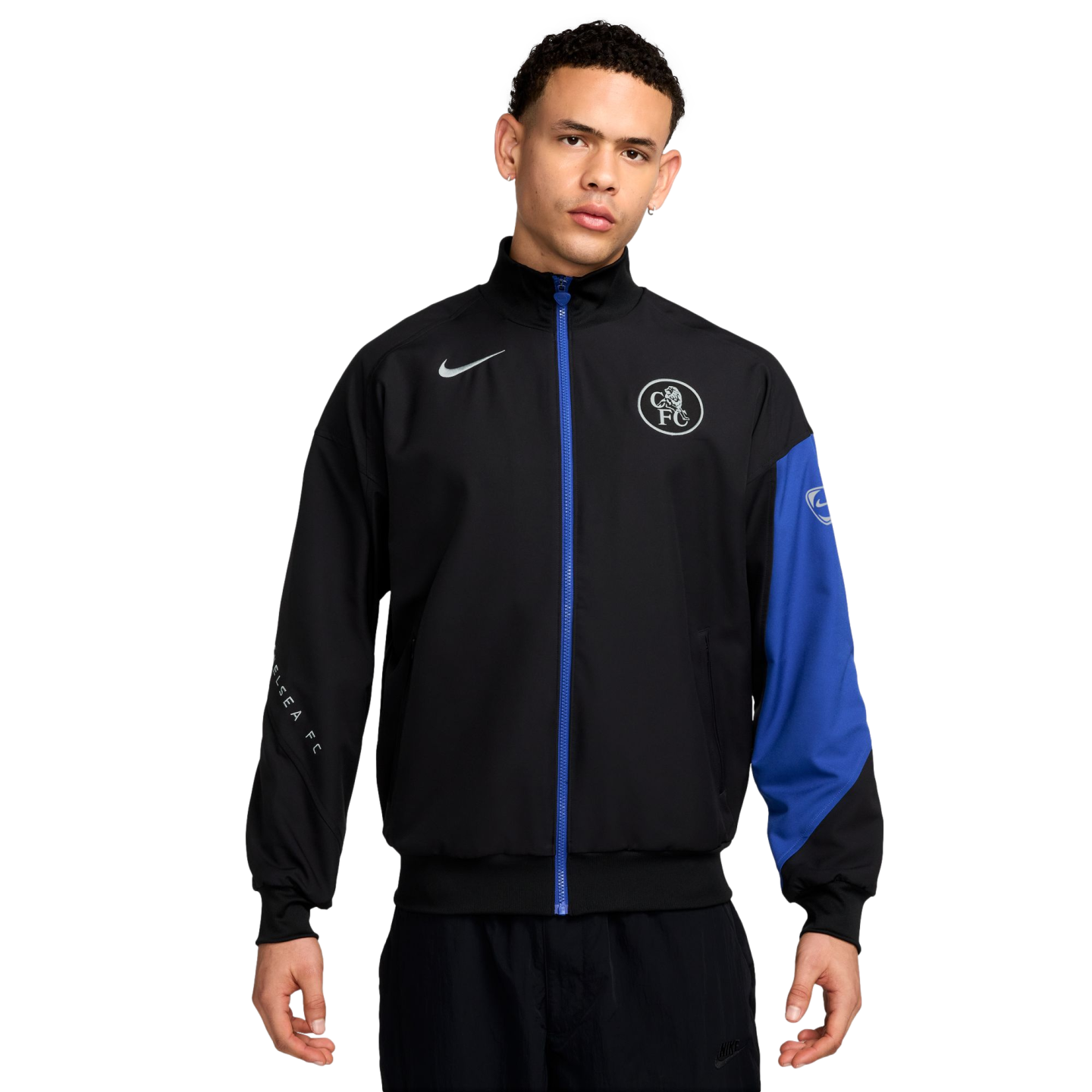 A man wearing a black Nike Chelsea FC track jacket with a blue zipper and white logos, featuring a blue panel on one sleeve, against a black background.