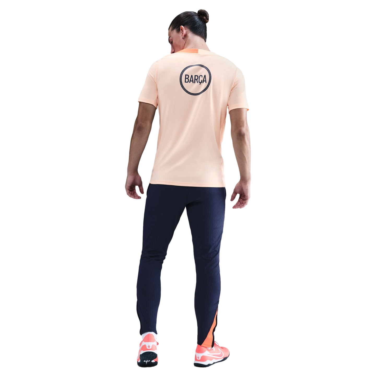Rear view of a man wearing a peach "BARÇA" t-shirt, navy joggers, and orange sneakers, set against a black background.