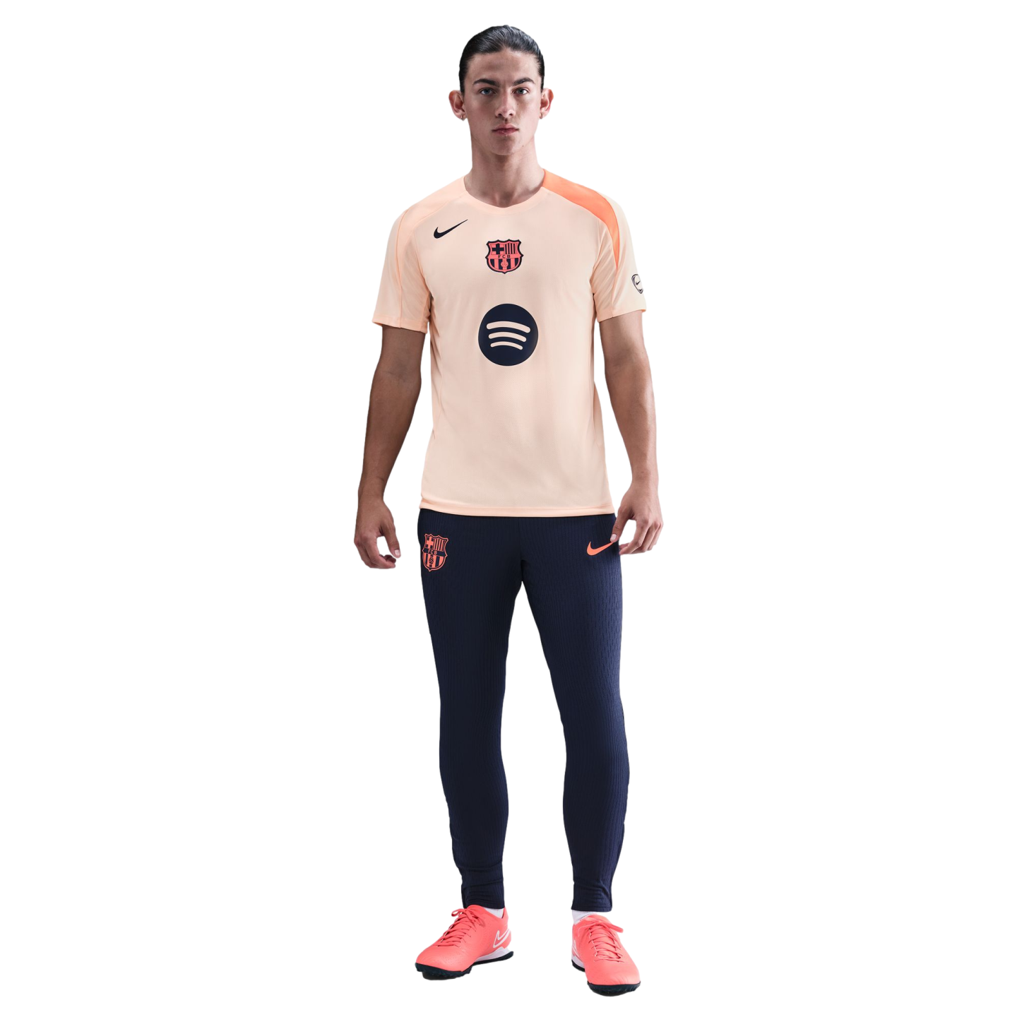 A full-length photo of a man wearing a peach FC Barcelona Nike training shirt, navy blue joggers, and coral soccer shoes against a black background.