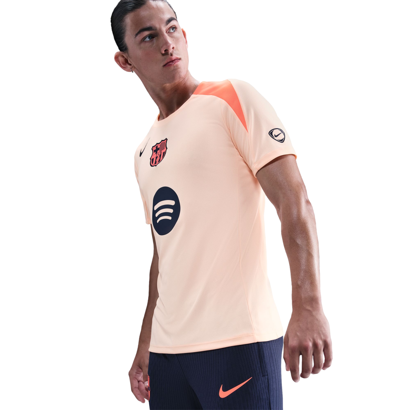 A man wearing a peach Nike FC Barcelona training top and navy shorts, looking off-camera against a black background.
