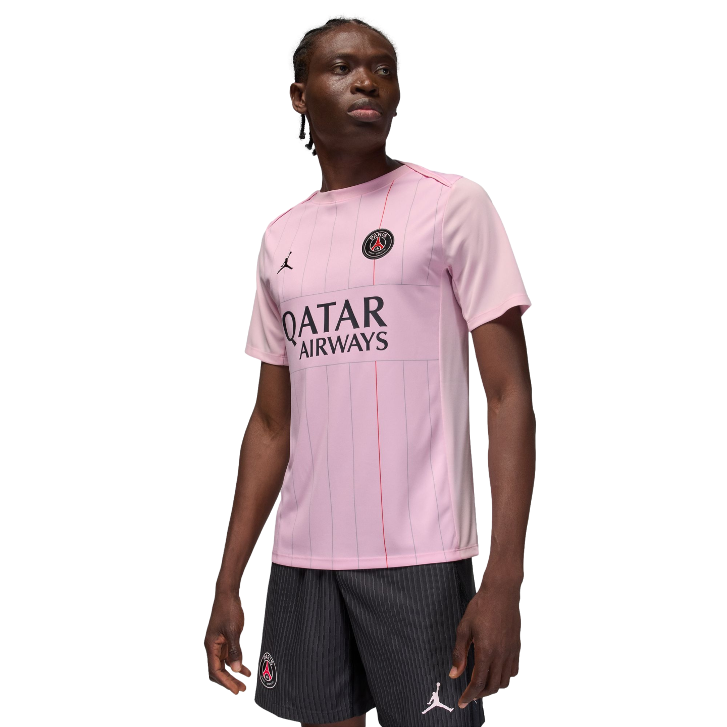 A man models a light pink Jordan Brand Paris Saint-Germain soccer jersey and matching black pinstripe shorts against a black background.