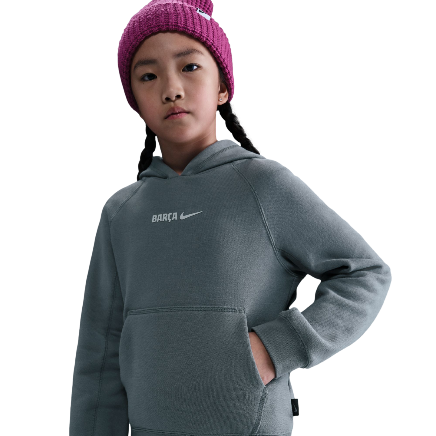 A young girl in a pink knit beanie and a grey Nike "Barça" hoodie looks forward against a solid black background.