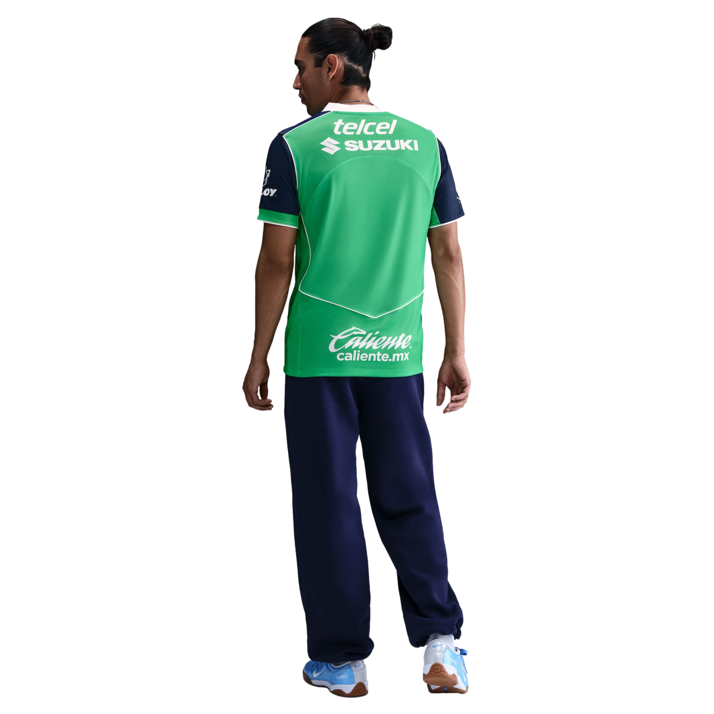 A man seen from behind wears a green and navy blue sports jersey with sponsor logos, navy pants, and light blue sneakers against a black background.