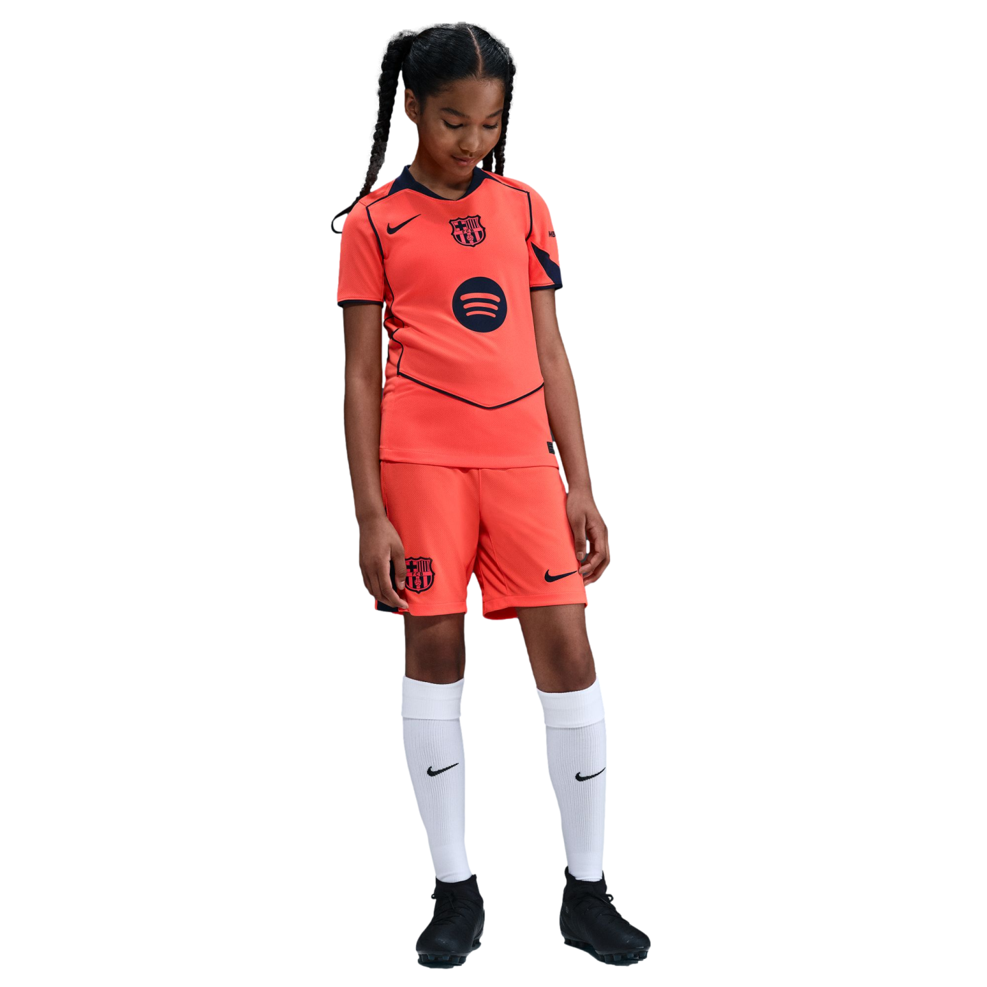 A young girl in a bright orange FC Barcelona soccer kit, white socks, and black cleats, standing against a solid black background.