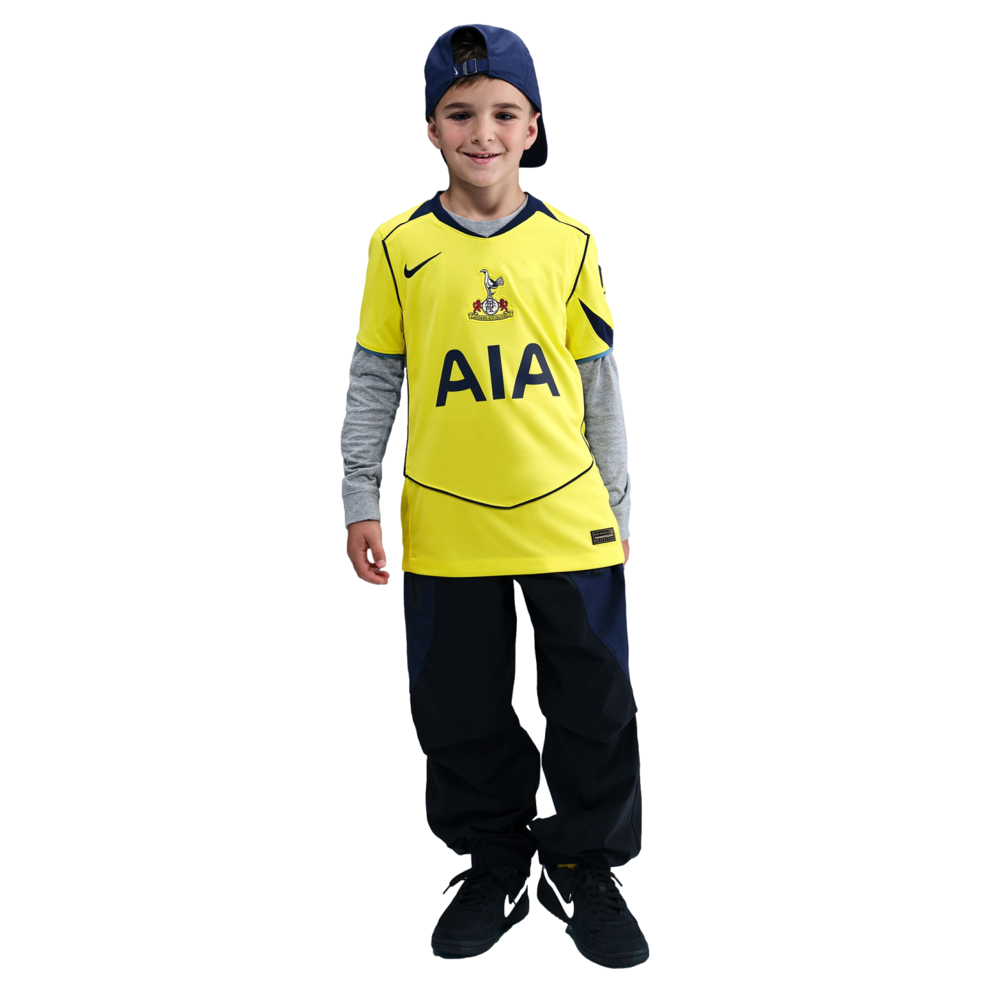 A smiling boy in a yellow Tottenham Hotspur jersey, grey long-sleeved shirt, navy cap, and dark pants, standing against a black background.
