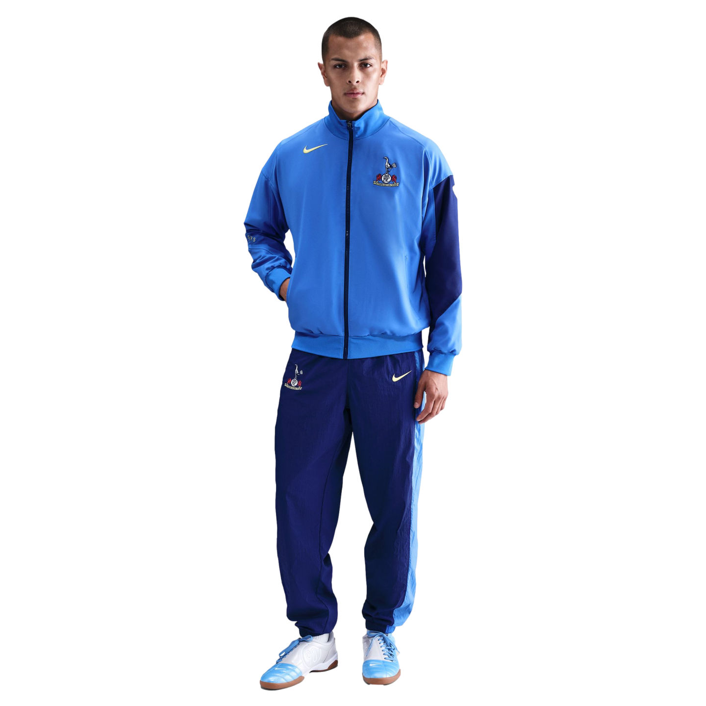 A man wearing a light blue and navy Nike Tottenham Hotspur FC tracksuit and matching sneakers, standing against a solid black background.