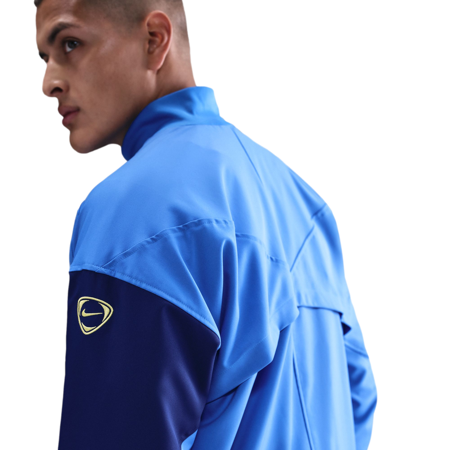 A man looking over his shoulder in a light blue and navy Nike jacket with a gold logo on the arm, set against a black background.