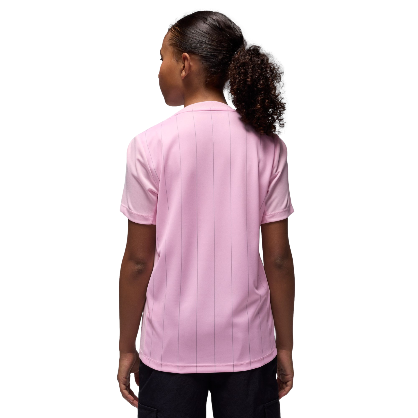 Rear view of a person with a curly ponytail wearing a light pink, pinstriped t-shirt and black shorts against a black background.