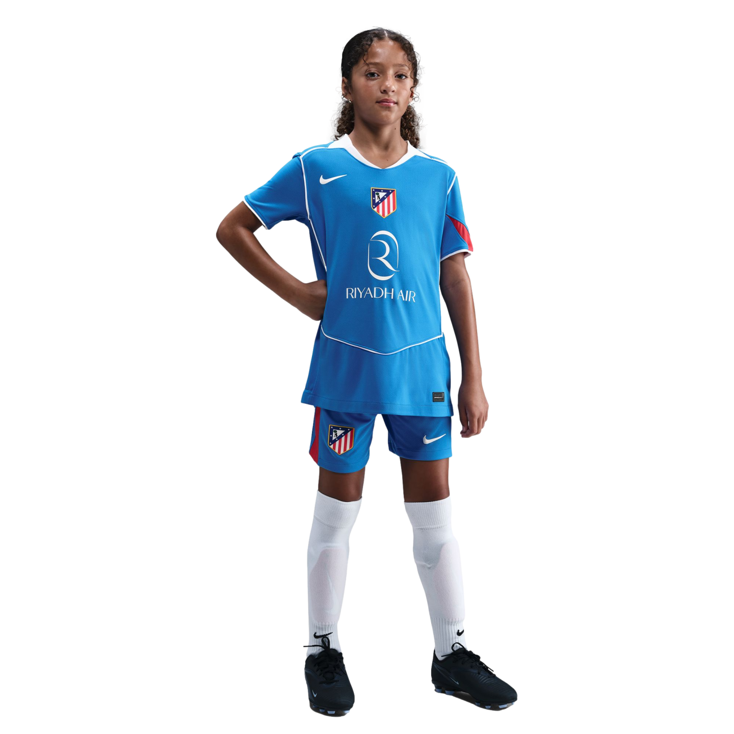 A young girl in a blue Atletico Madrid soccer kit with white socks and black cleats, posing with a hand on her hip against a black background.