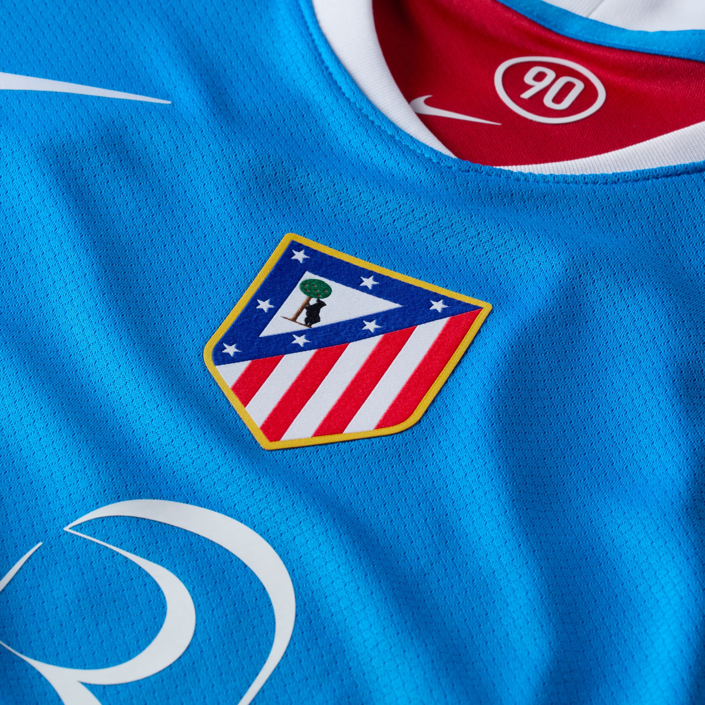 Close-up of a blue Nike athletic jersey featuring the Atlético Madrid crest and a red inner collar with a circular "90" logo.