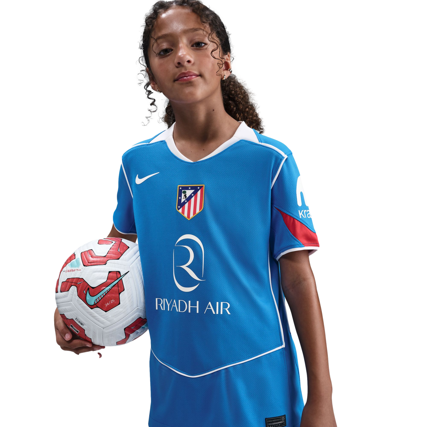 A young girl with curly hair poses in a blue Atlético Madrid soccer jersey, holding a soccer ball under her arm against a black background.