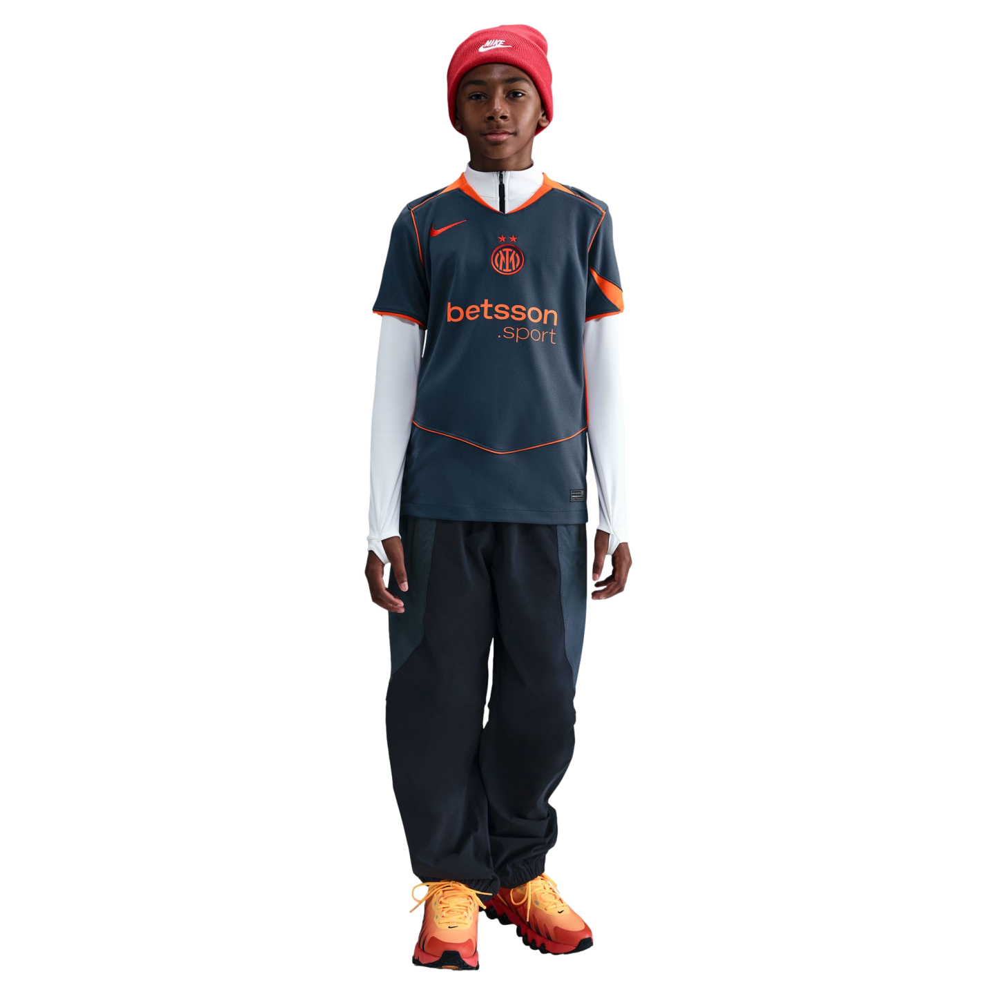 Young person in a red Nike beanie, grey and orange Inter Milan jersey, dark pants, and orange sneakers standing against a black background.