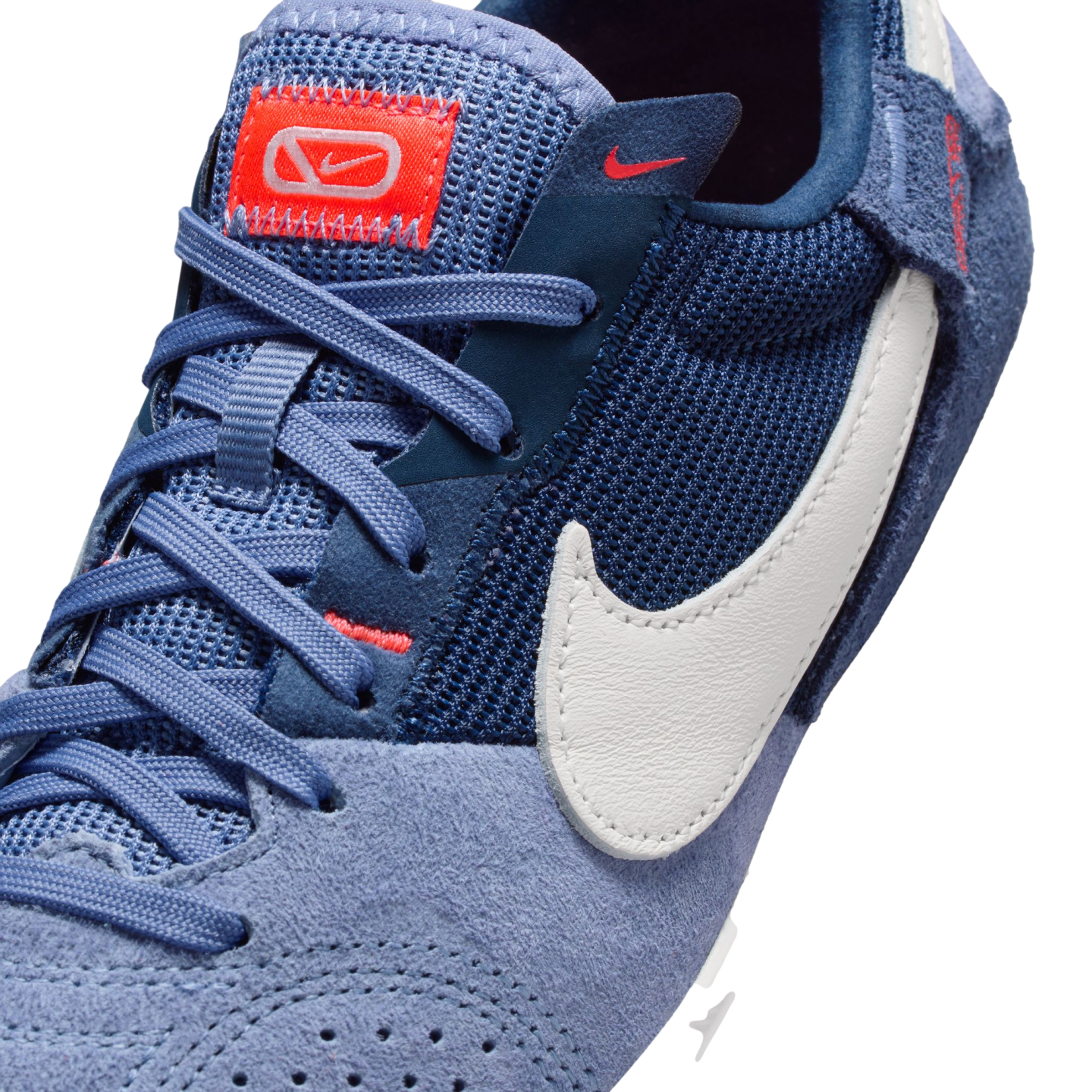 Close-up of a blue Nike sneaker featuring suede and mesh materials, light blue laces, a white swoosh logo, and an orange brand tag on the tongue.