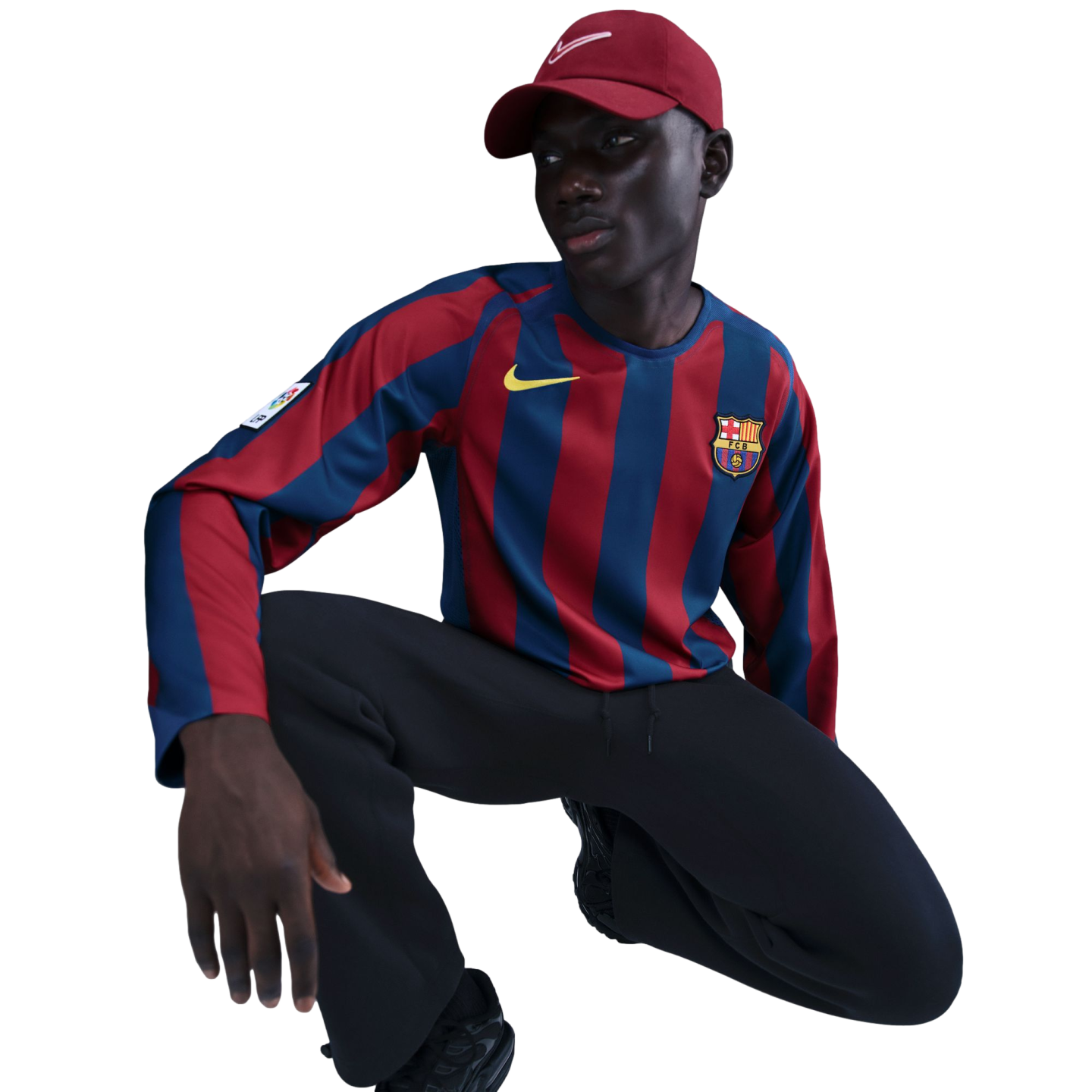 A person in a long-sleeved FC Barcelona jersey, dark pants, and a maroon Nike cap crouches against a solid black background.