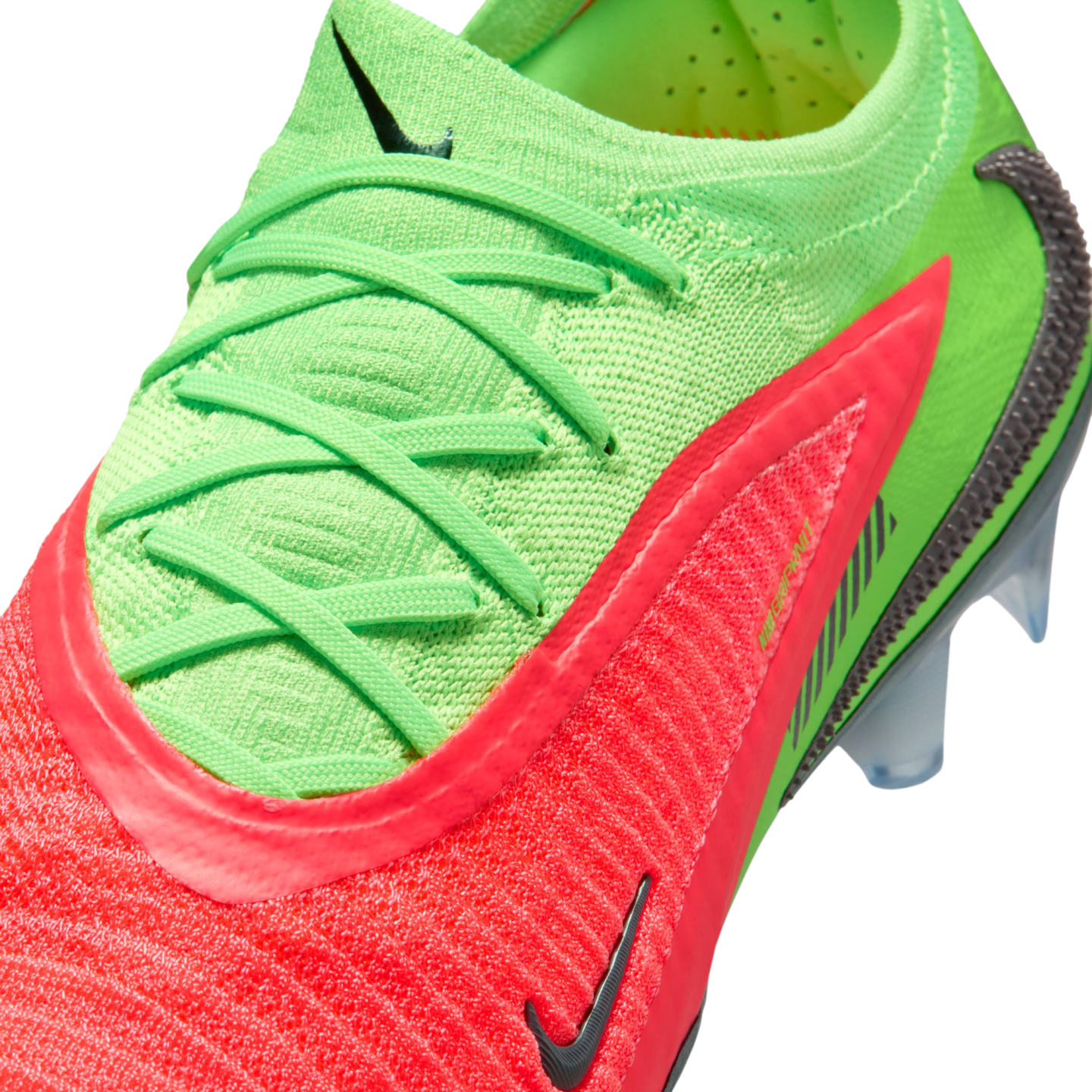 A close-up, high-angle view of a vibrant neon green and bright pink Nike soccer cleat against a black background. The shoe features a textured, knitted upper with matching neon green laces and multiple black Nike swoosh logos. Clear, translucent studs are