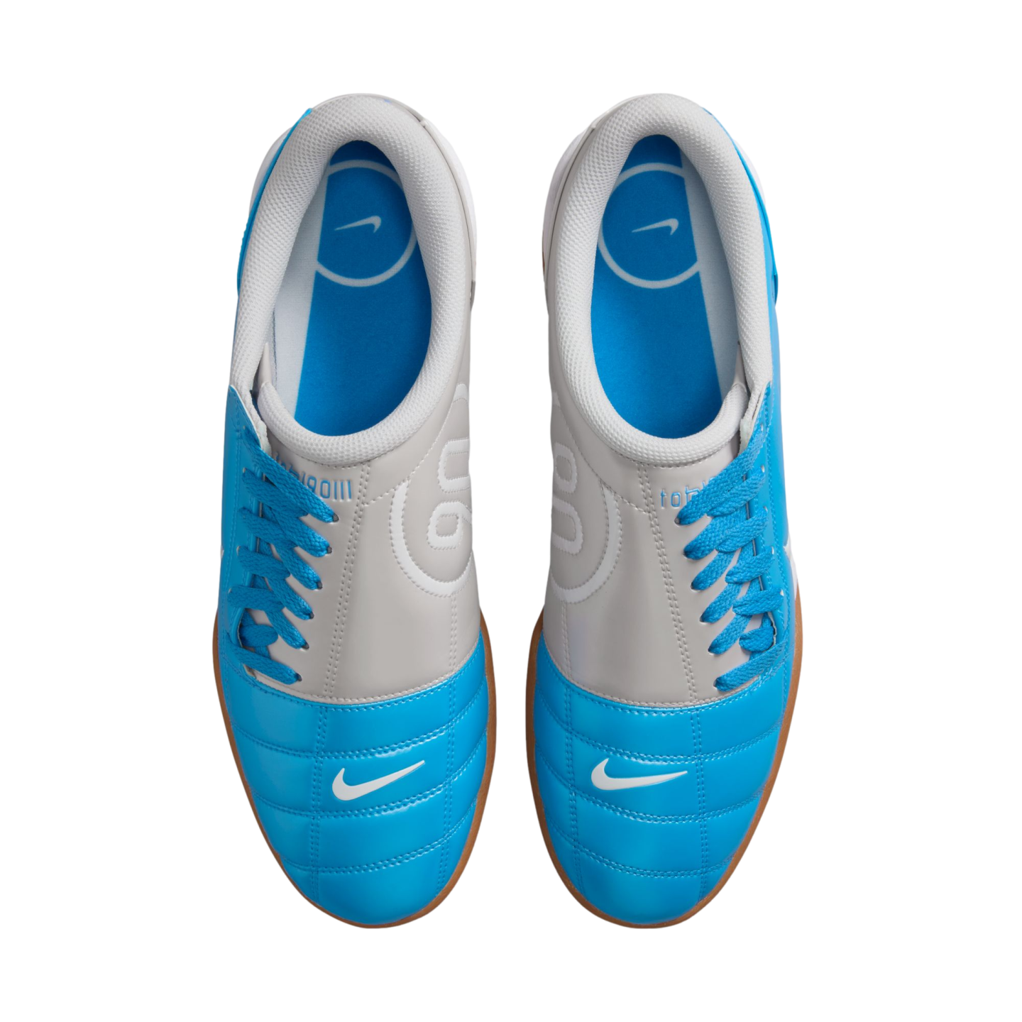 Top-down view of blue and grey Nike indoor soccer shoes with white swooshes and blue laces, resting on a black background.