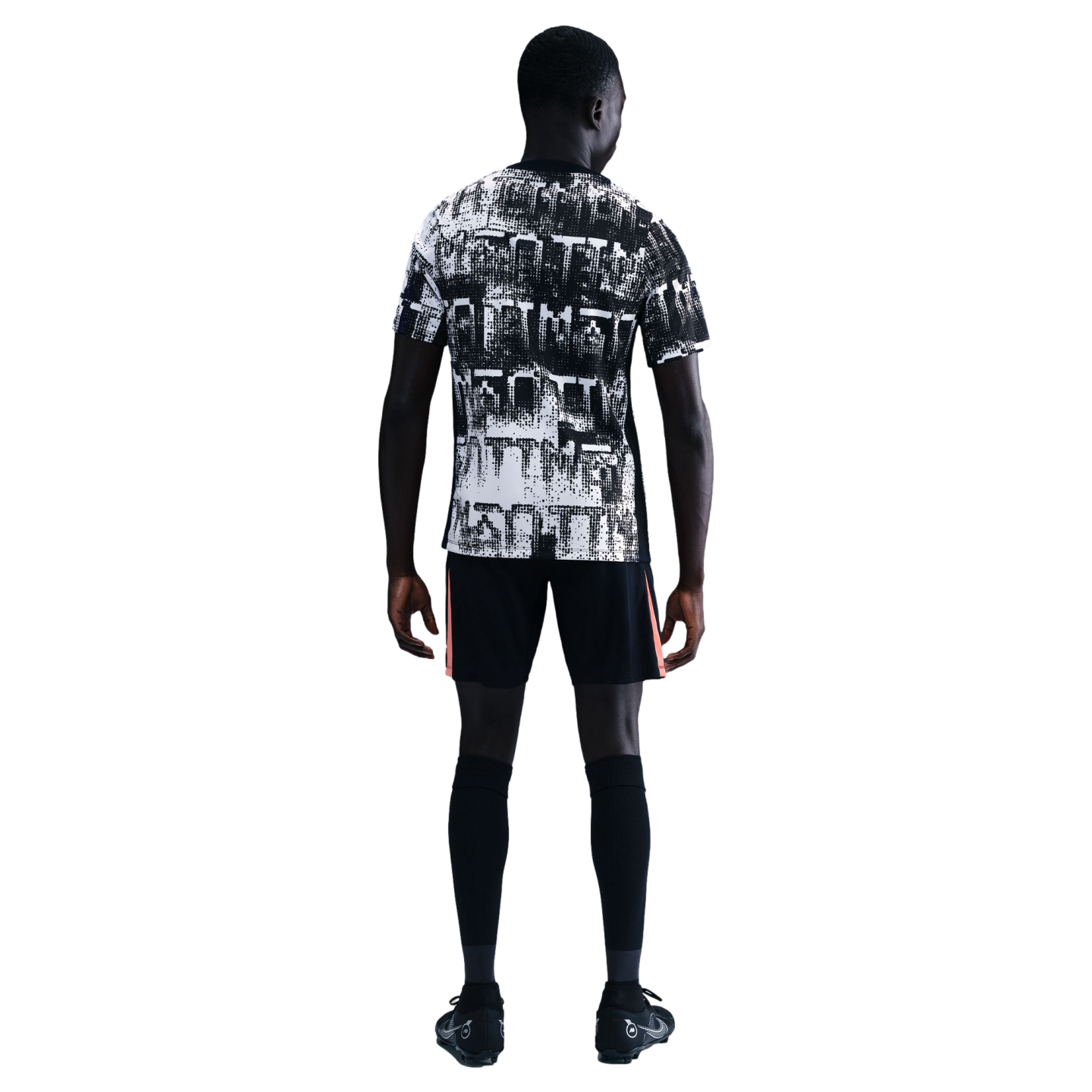 Rear view of a man in a black and white graphic t-shirt, black shorts with orange details, and high socks against a solid black background.