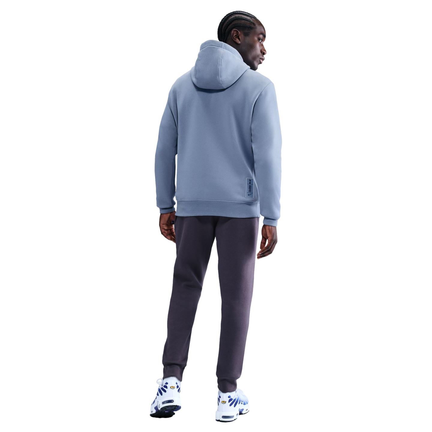 Rear view of a man with braids wearing a light blue hoodie and dark grey joggers against a black background.