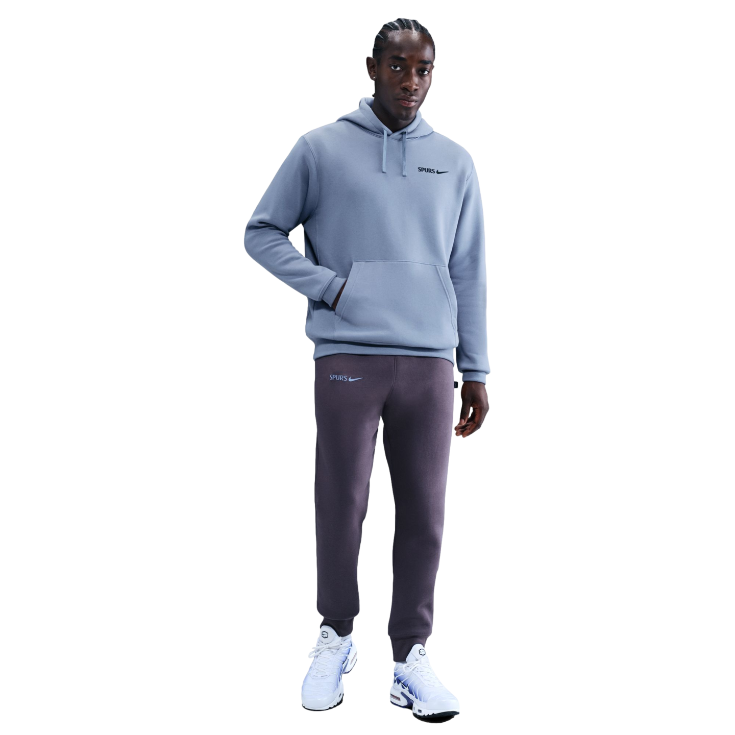 A man models a light blue Nike Spurs hoodie, dark purple sweatpants, and white sneakers against a black background.