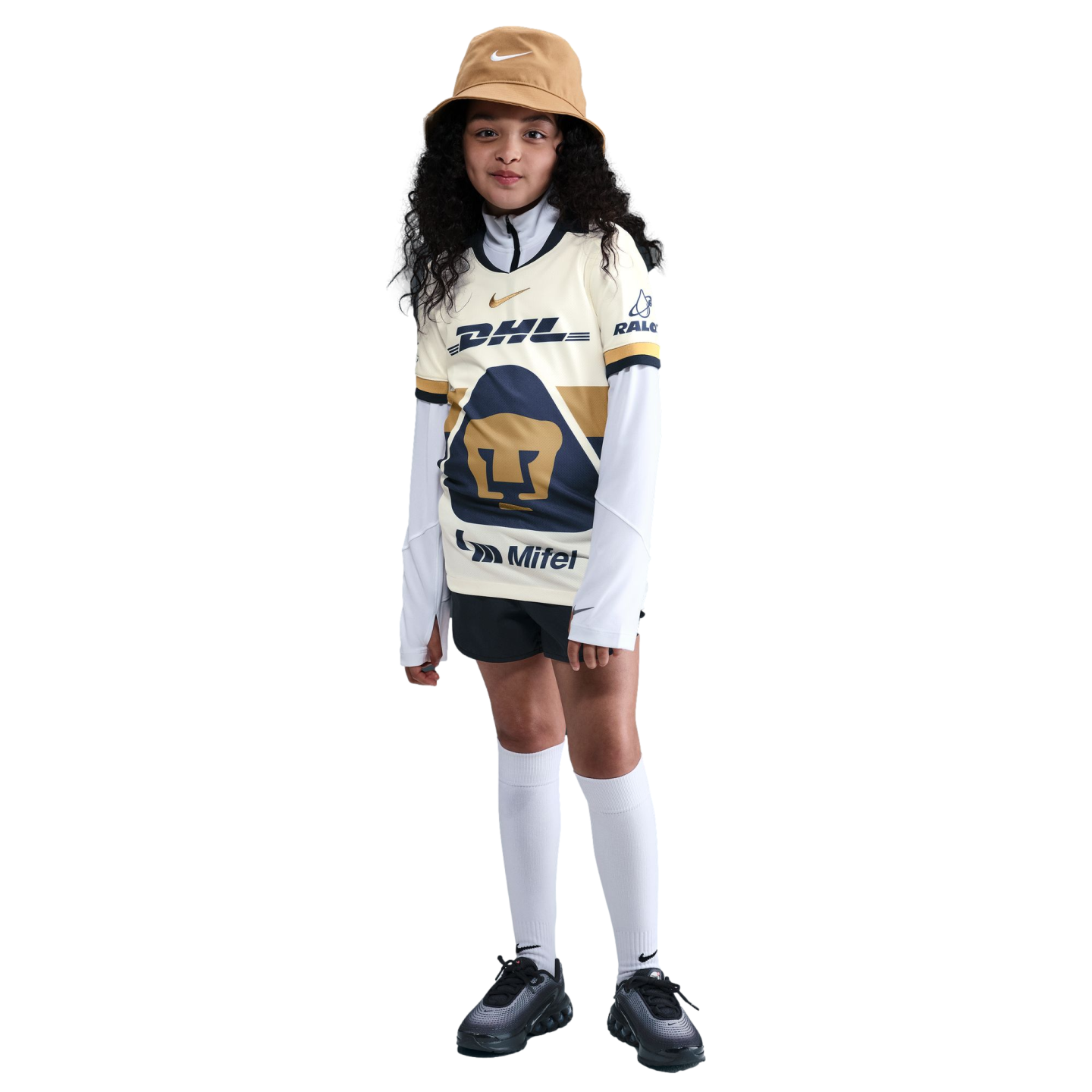 A young girl with long curly hair wearing a soccer jersey, tan bucket hat, black shorts, and white knee-high socks, standing against a plain white background.