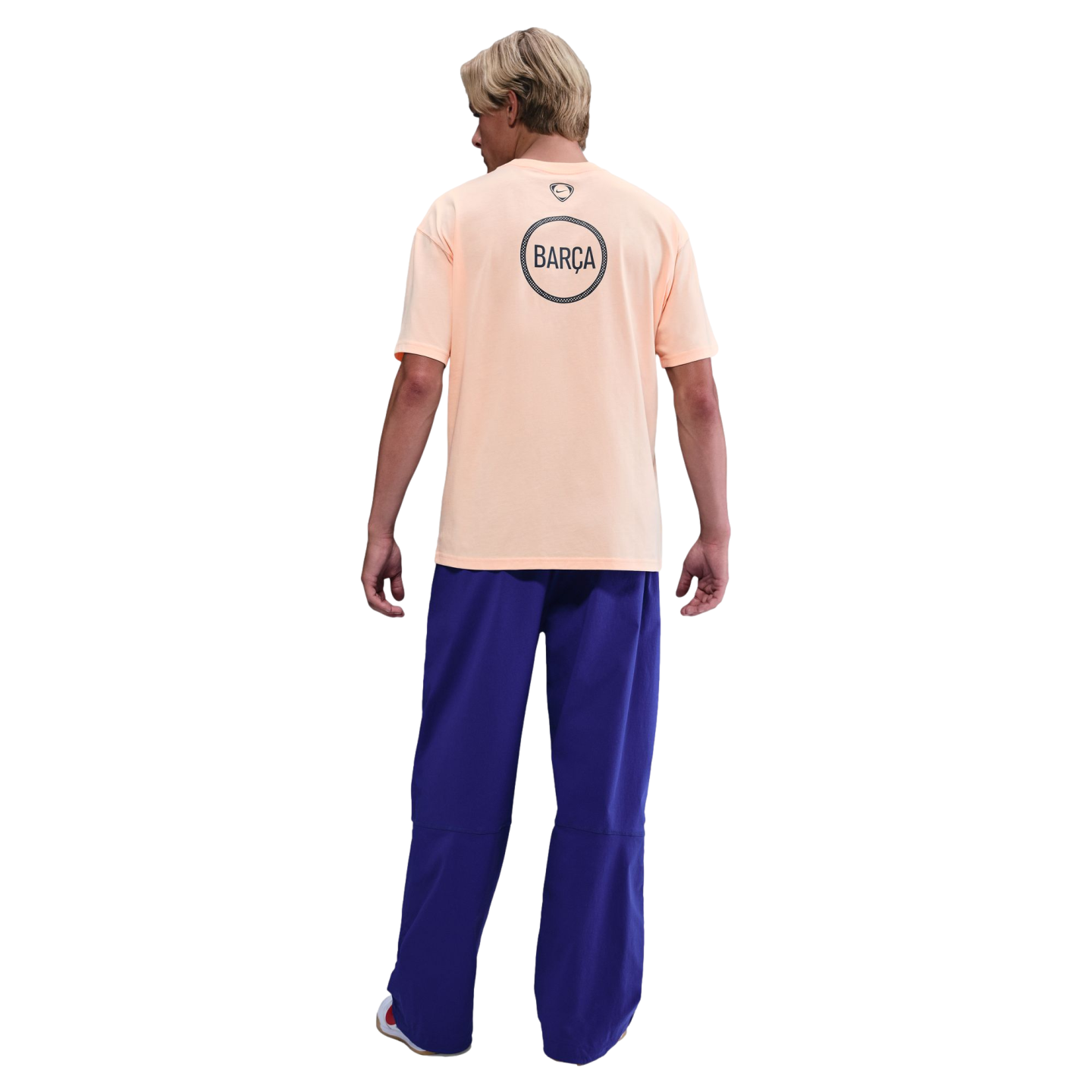 Back view of a man wearing a peach-colored Nike Barça t-shirt and baggy blue pants against a black background.