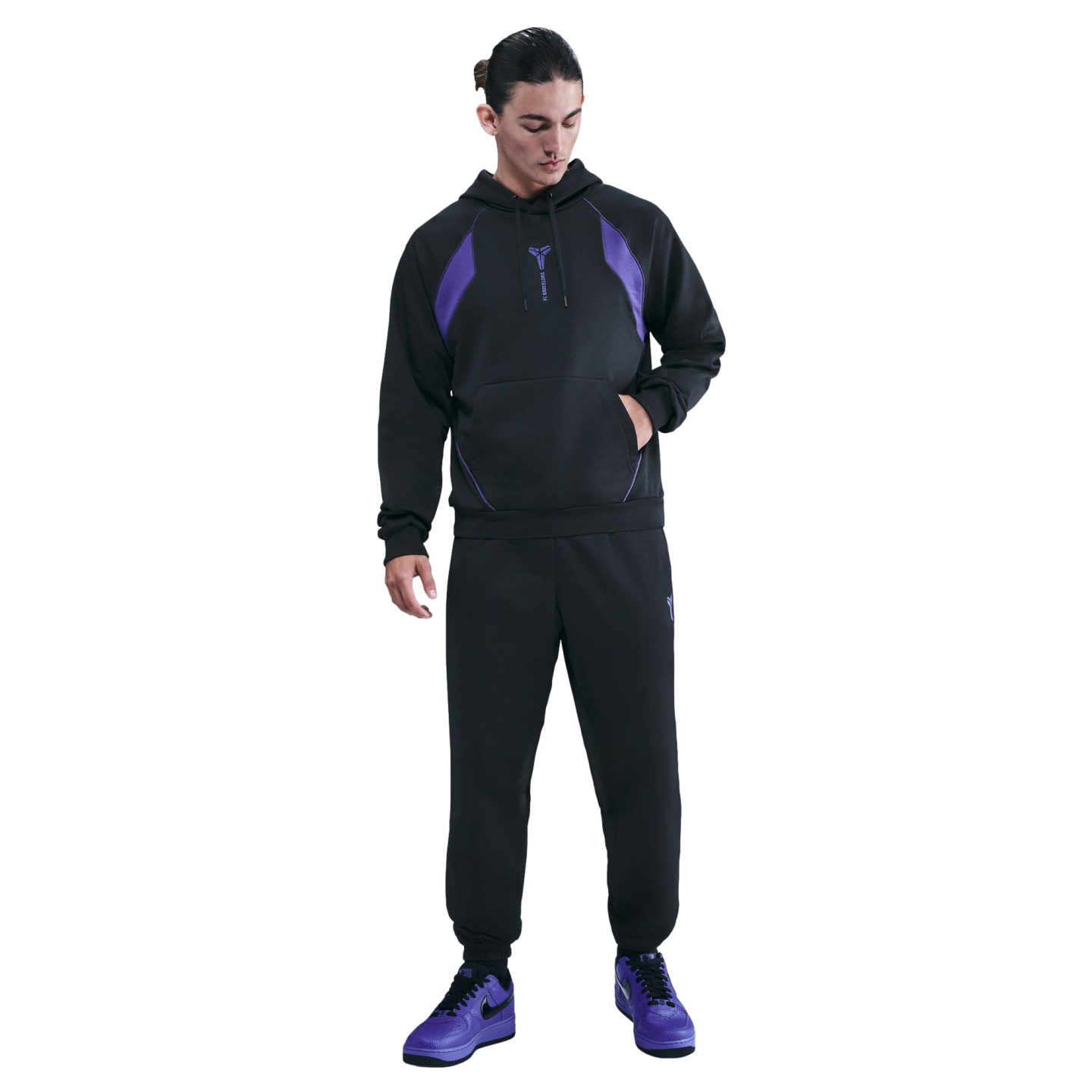 A man wears a black and purple Nike Kobe Bryant x FC Barcelona tracksuit and purple sneakers, standing against a black background.