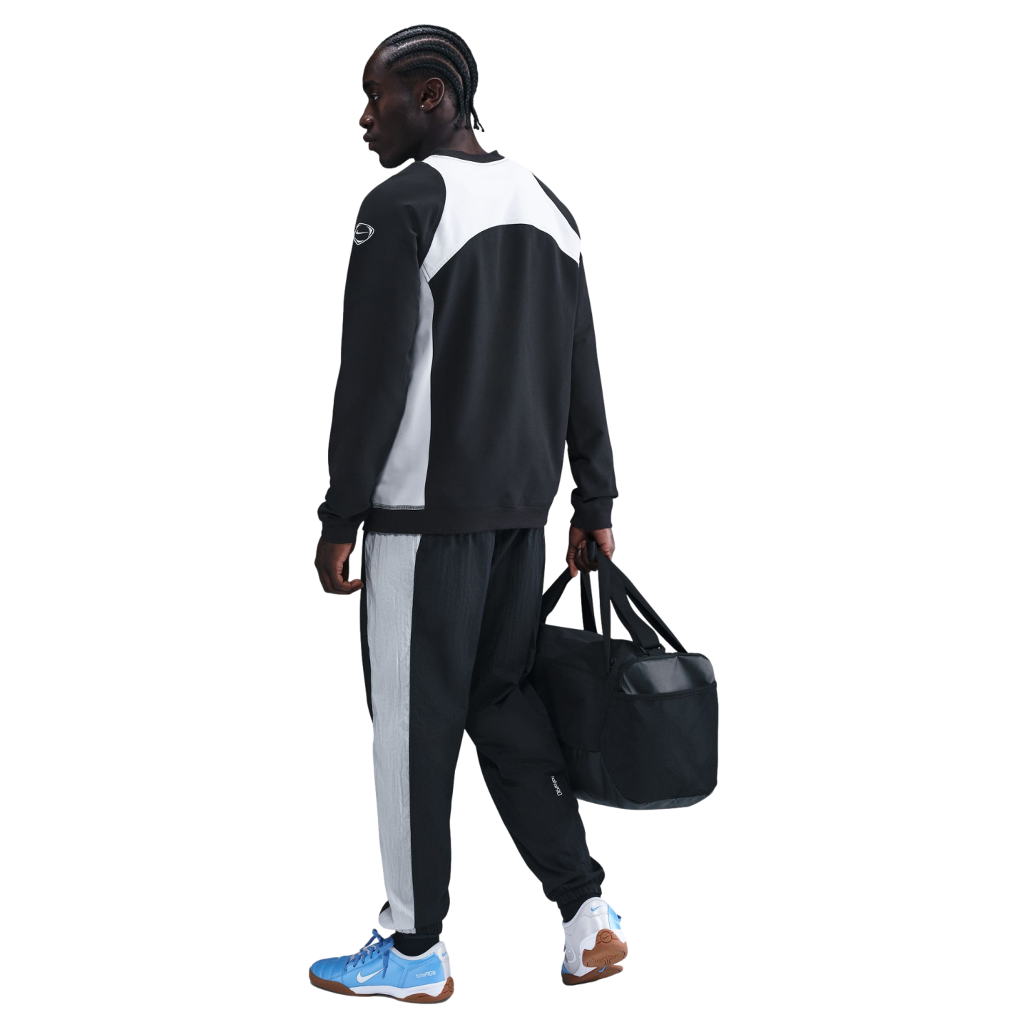 A person walks away wearing a black and white Nike tracksuit and light blue sneakers, carrying a black duffel bag against a solid black background.
