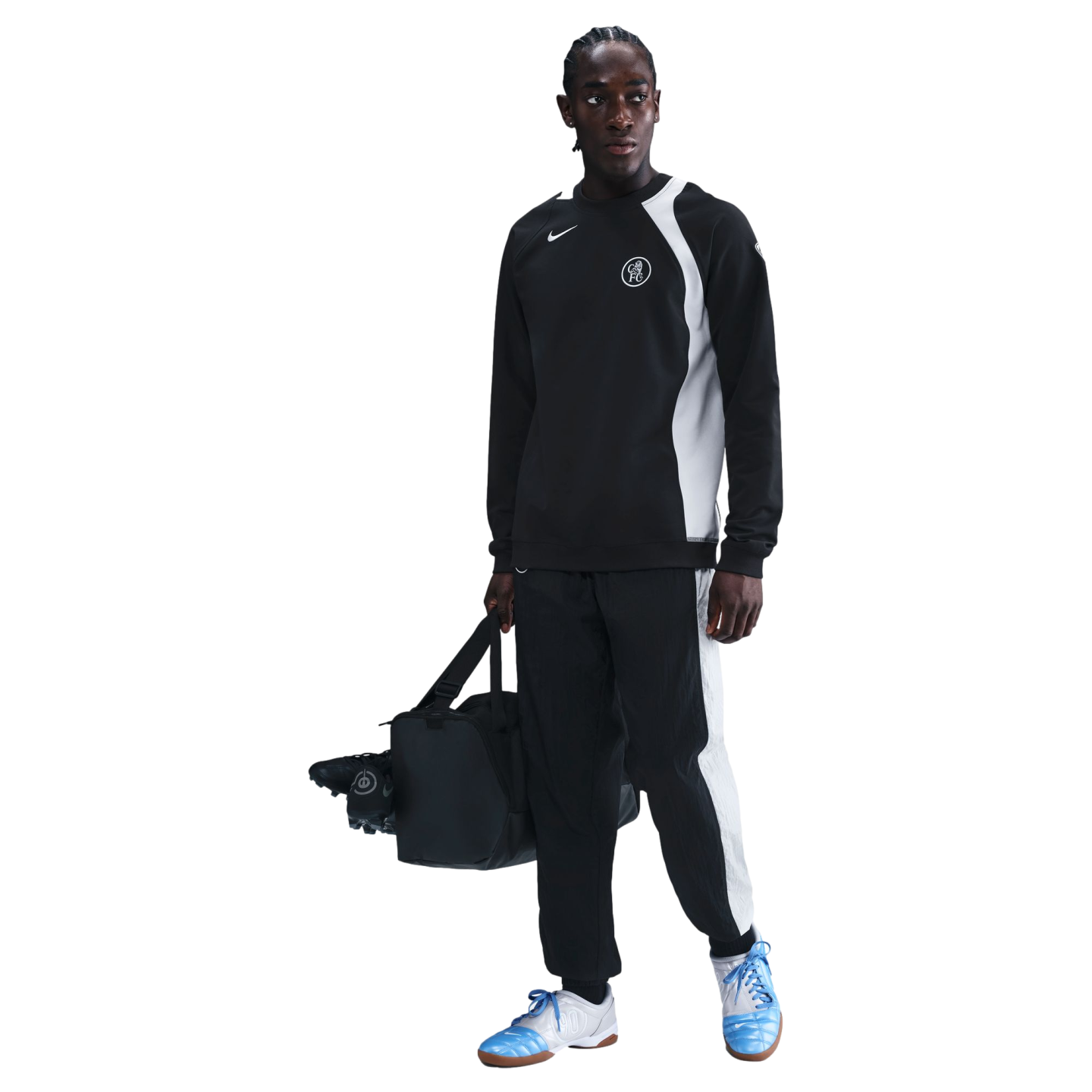 A man in a black Chelsea FC Nike tracksuit holding a duffel bag and soccer cleats, wearing light blue sneakers against a black background.