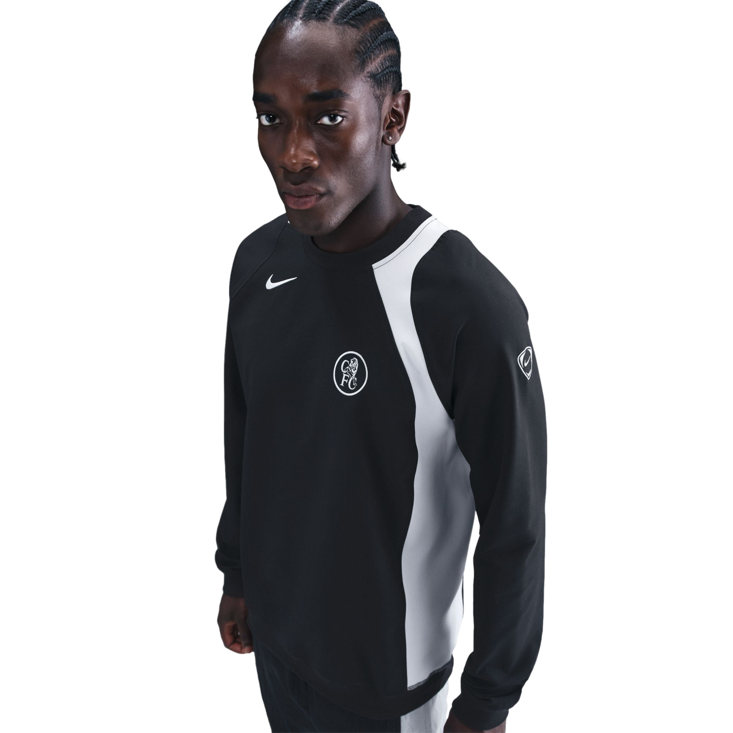 A man with braided hair wearing a black Nike sweatshirt with a white side panel and Chelsea FC logo, posed against a solid black background.