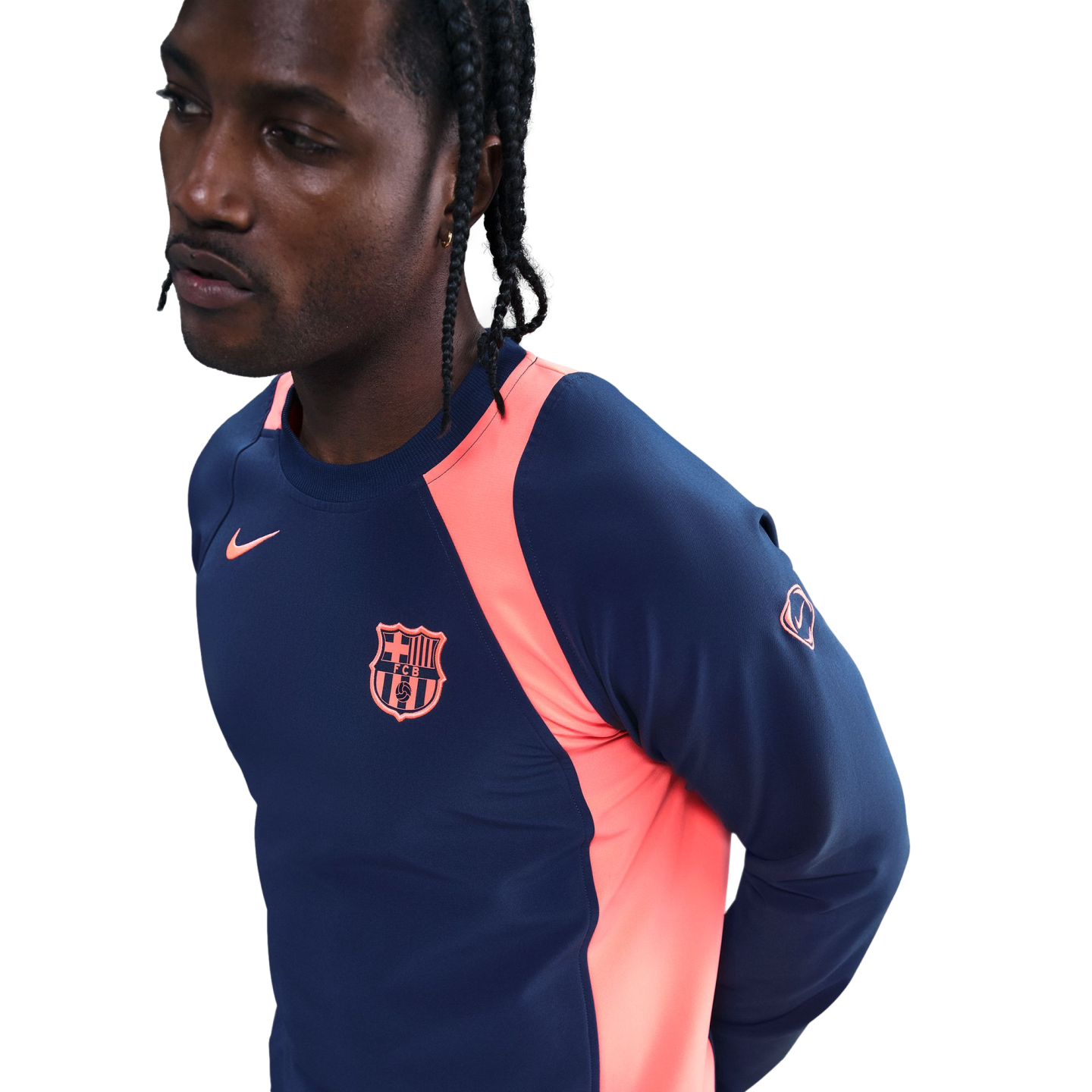 Man with braids wearing a navy and pink Nike FC Barcelona long-sleeve training shirt, looking away against a black background.