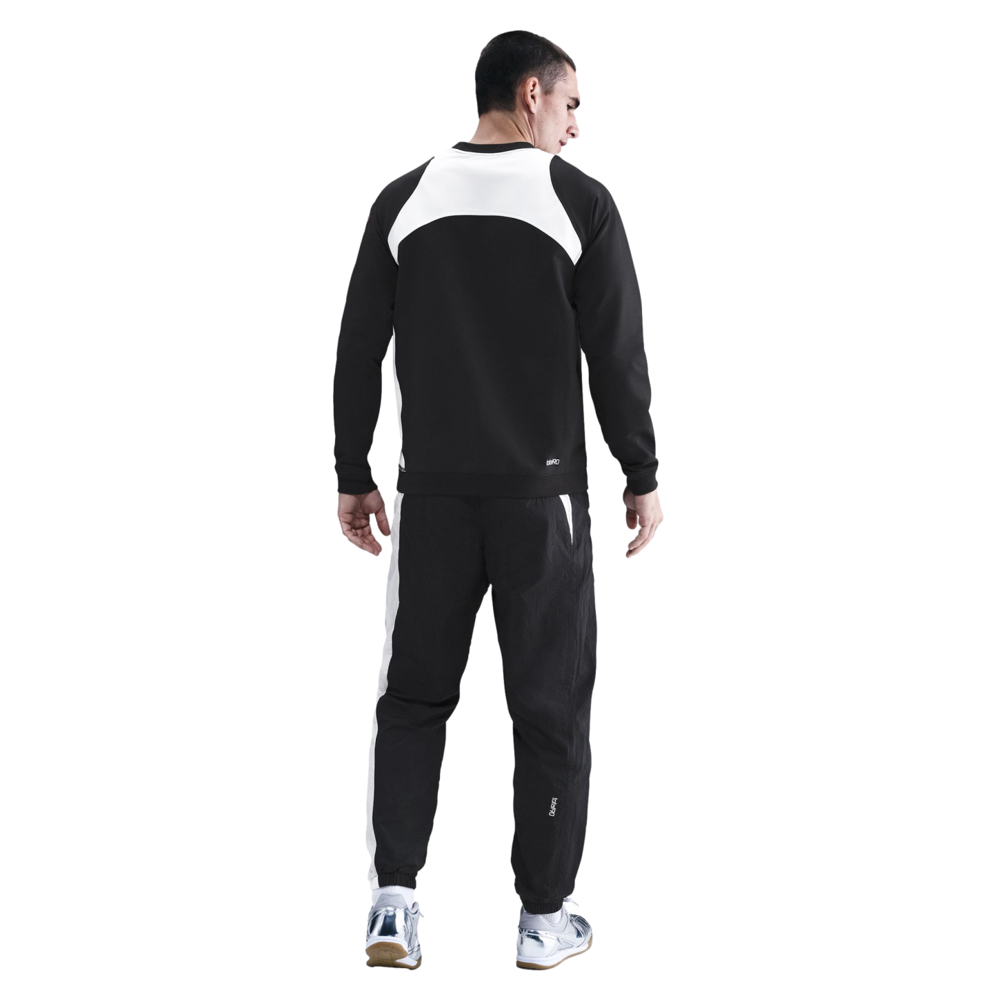 Rear view of a man wearing a black and white tracksuit and silver sneakers against a black background.