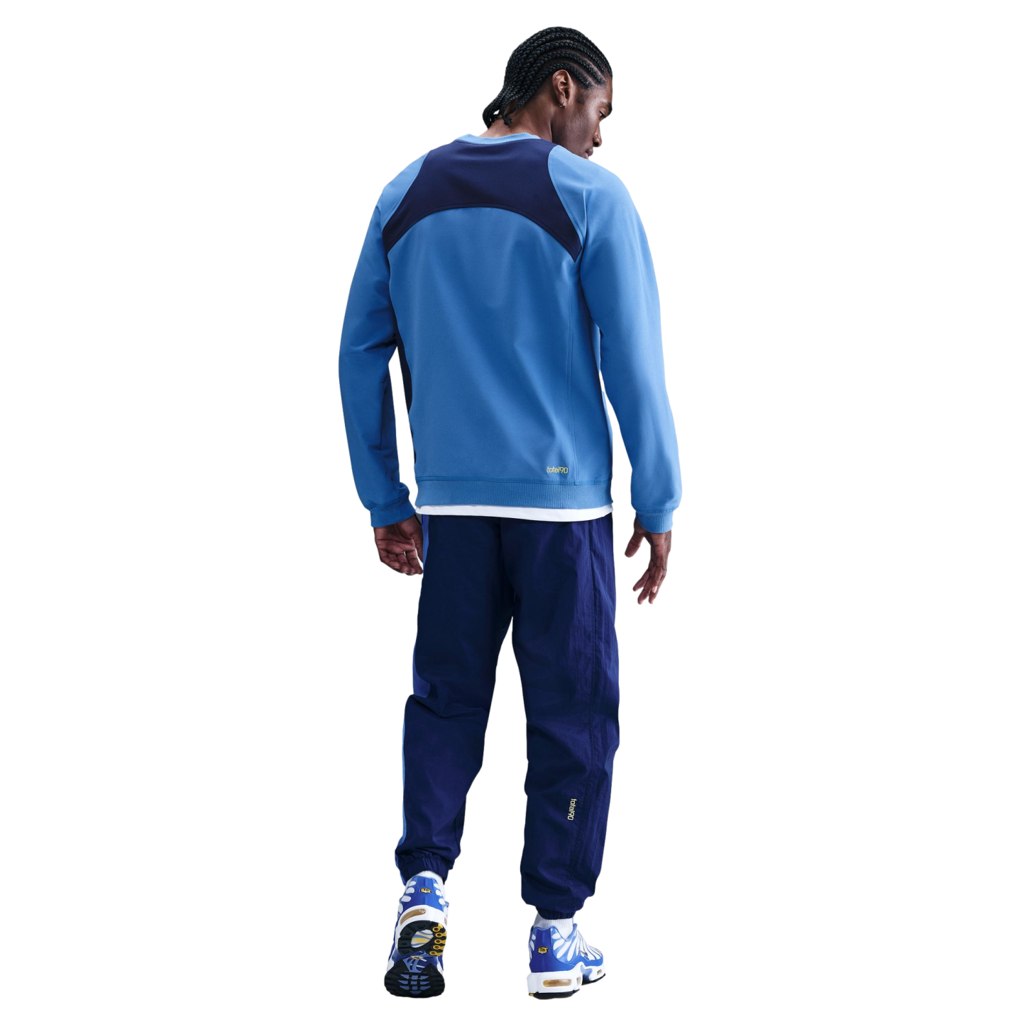 Back view of a man with braided hair wearing a blue and navy athletic tracksuit and sneakers against a solid black background.