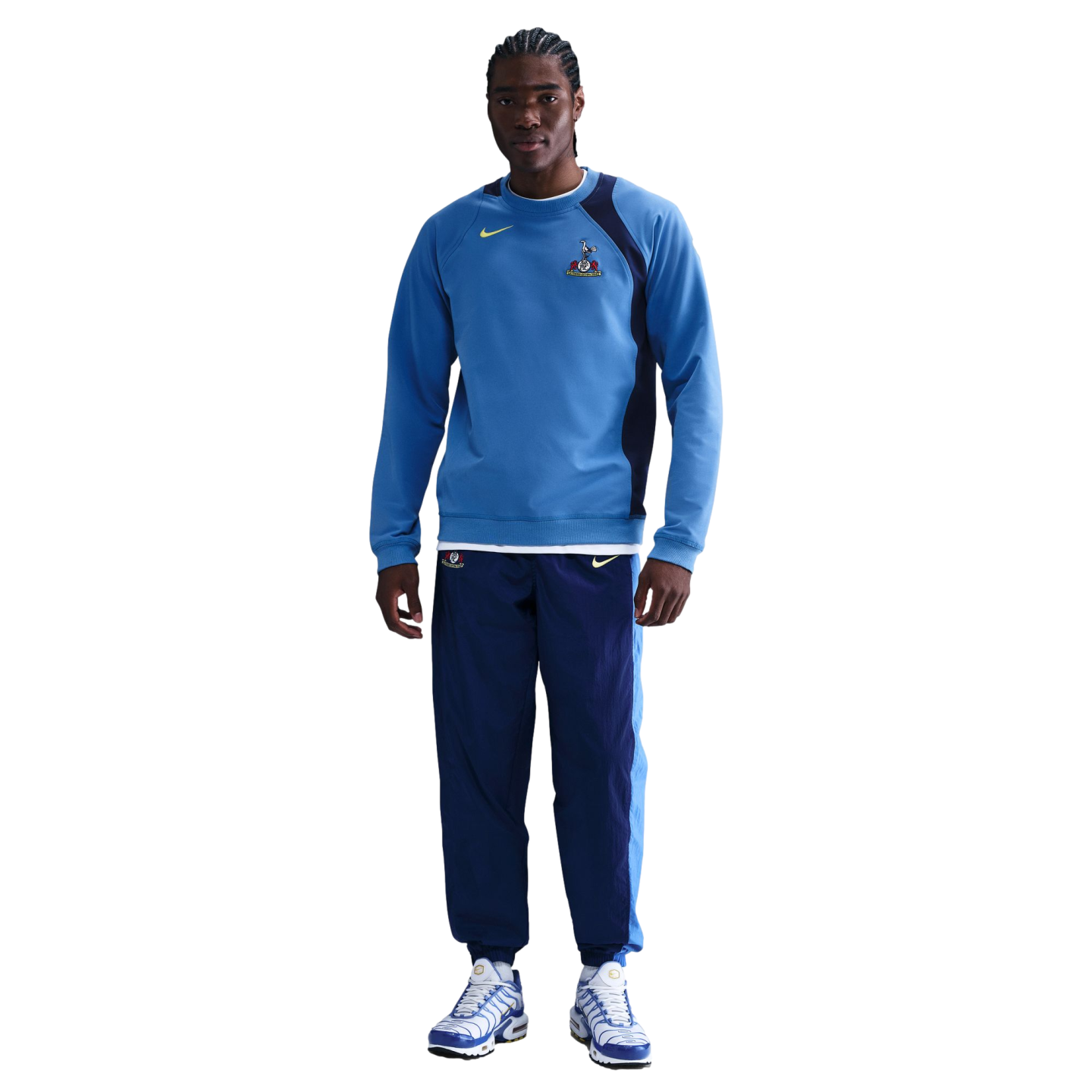 Man wearing a blue Tottenham Hotspur Nike sweatshirt, navy track pants, and blue and white sneakers against a black background.