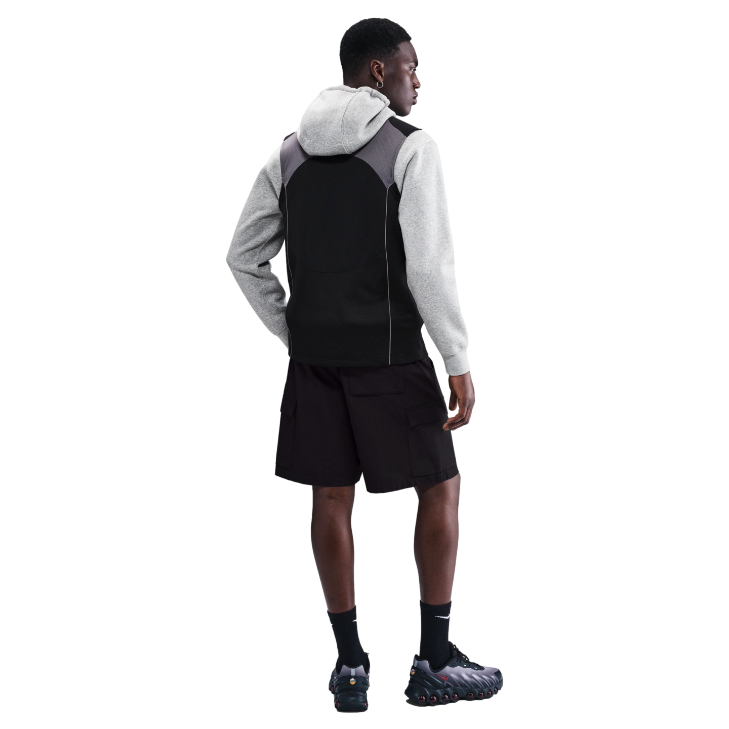 Rear view of a man in a grey hoodie, black vest, black cargo shorts, and sneakers against a black background.